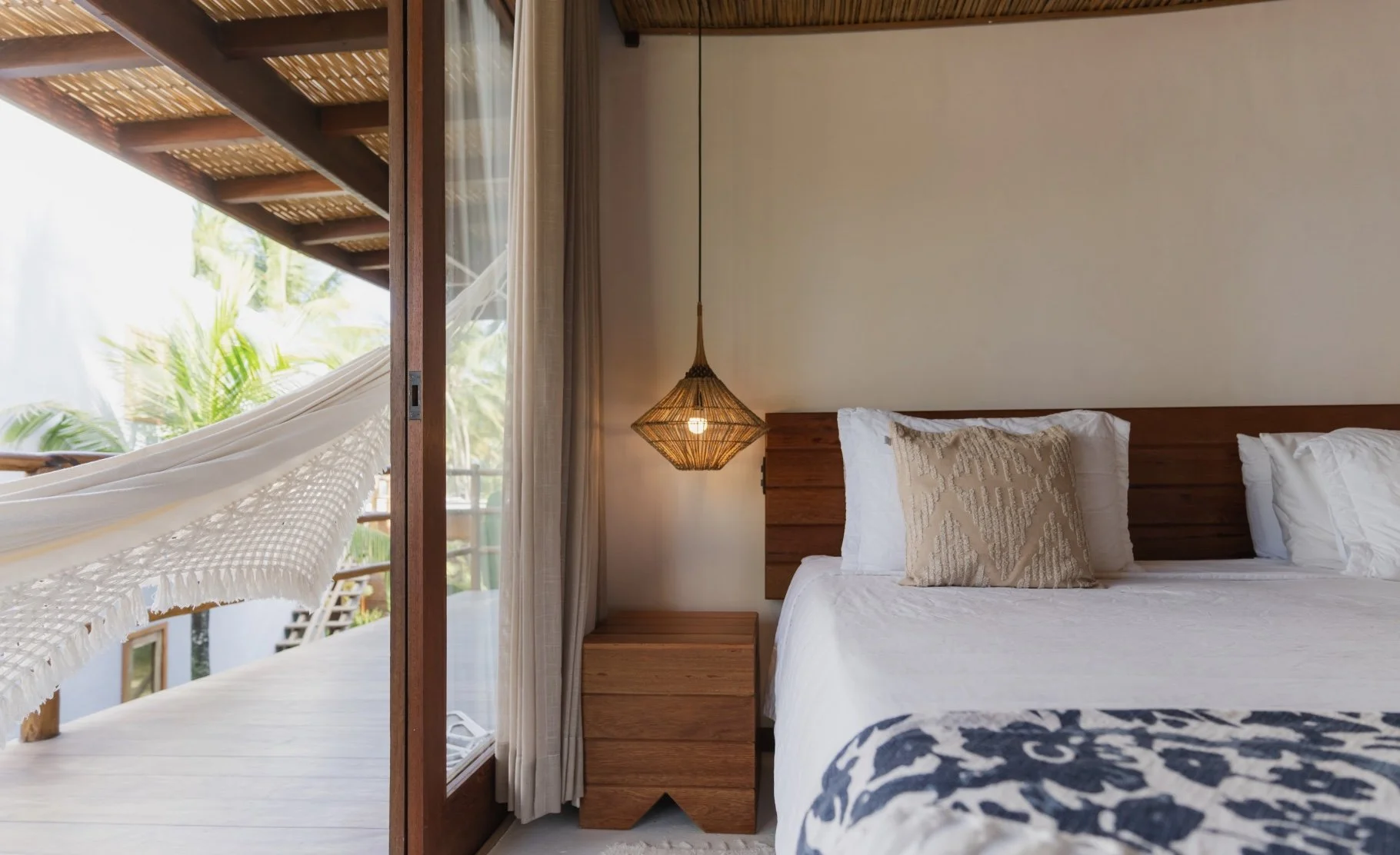Quarto de hotel com cama de casal, almofadas bege, cabeceira de madeira, luminária de fibra natural, cortinas claras, varanda com rede e plantas ao fundo.