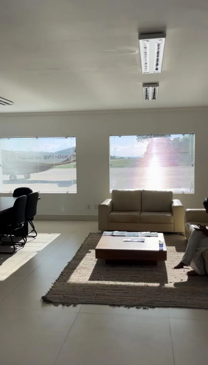 Sala de espera com sofá bege, mesa de centro de madeira, tapete de linho, duas janelas mostrando um avião na pista de aeroportos, e uma pessoa sentada em uma poltrona lendo algo.