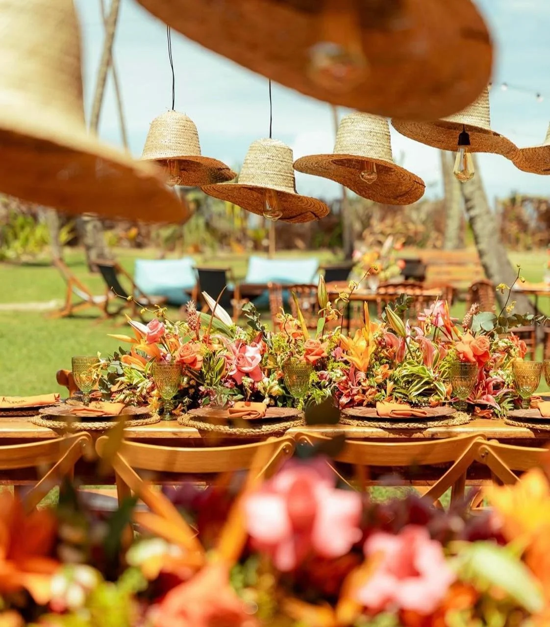 Decoração de mesa ao ar livre com arranjo de flores, copos e pratos, com chapéus de palha pendurados decorativamente acima.
