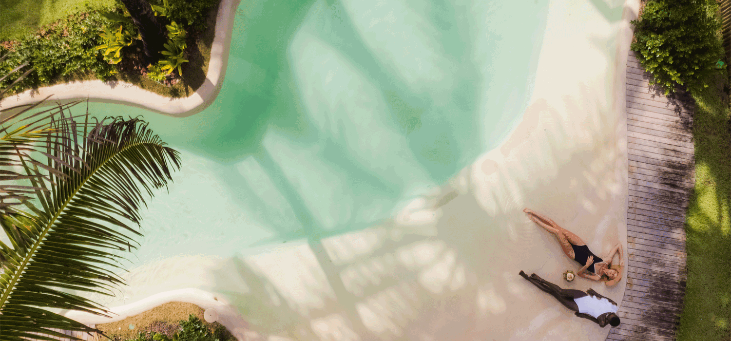 Pessoa deitada na praia com dois outros indivíduos ao lado, cercada por vegetação tropical e uma piscina