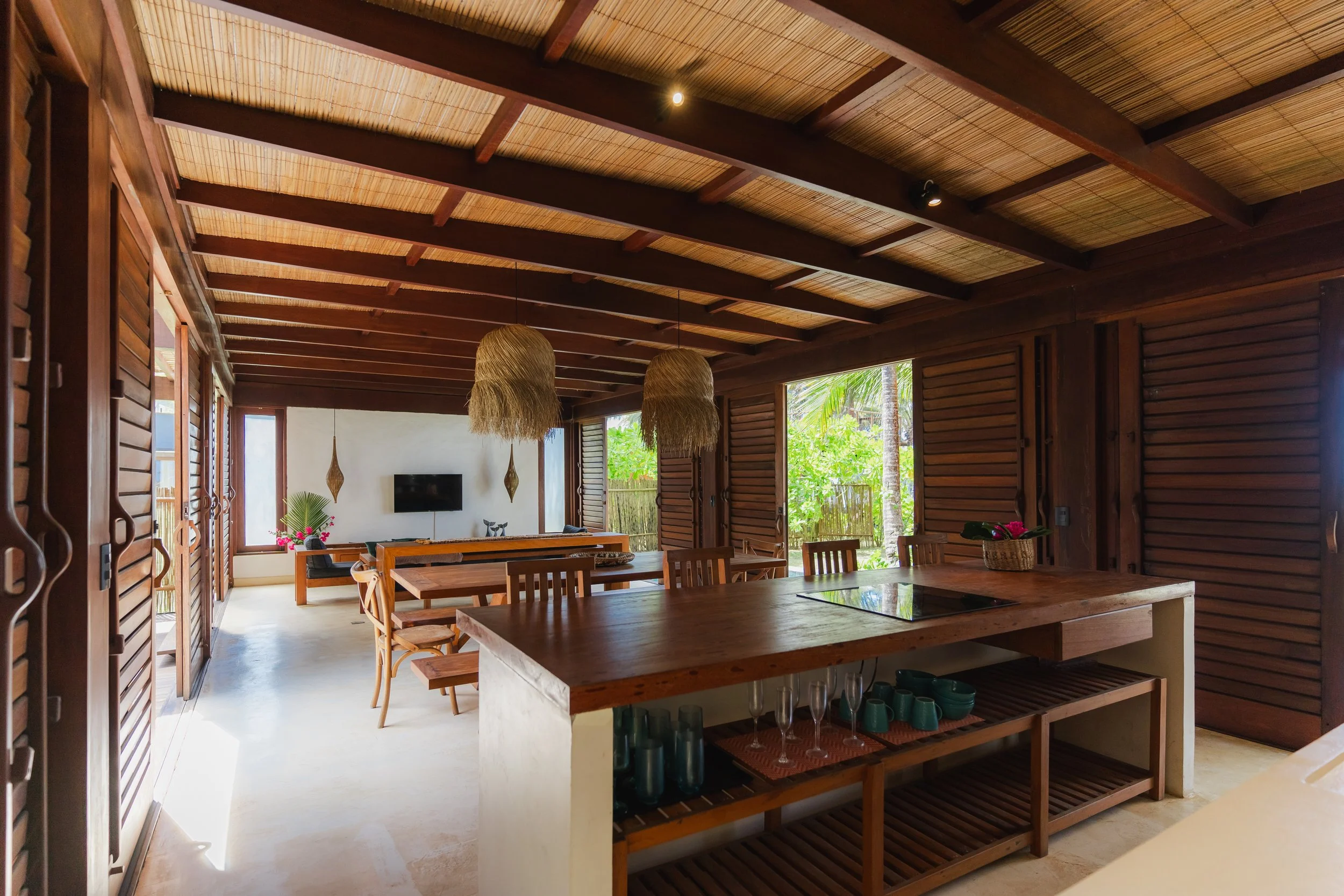 Interior de uma sala com teto de madeira, apoiado por vigas expostas, com iluminação embutida e luminárias de fibra natural. Possui janelas grandes com vistas para um jardim tropical, incluindo árvores verdes. Há uma mesa principal de madeira com cad