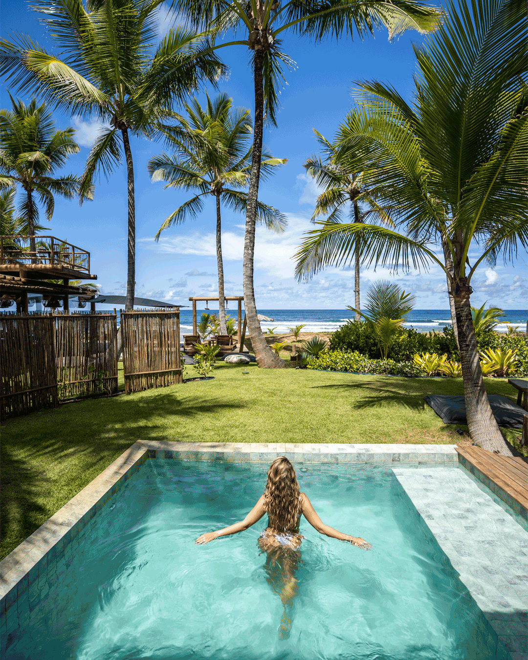 Pessoa nadando na piscina em um ambiente tropical com palmeiras, gramado, praia, oceano e céu azul.