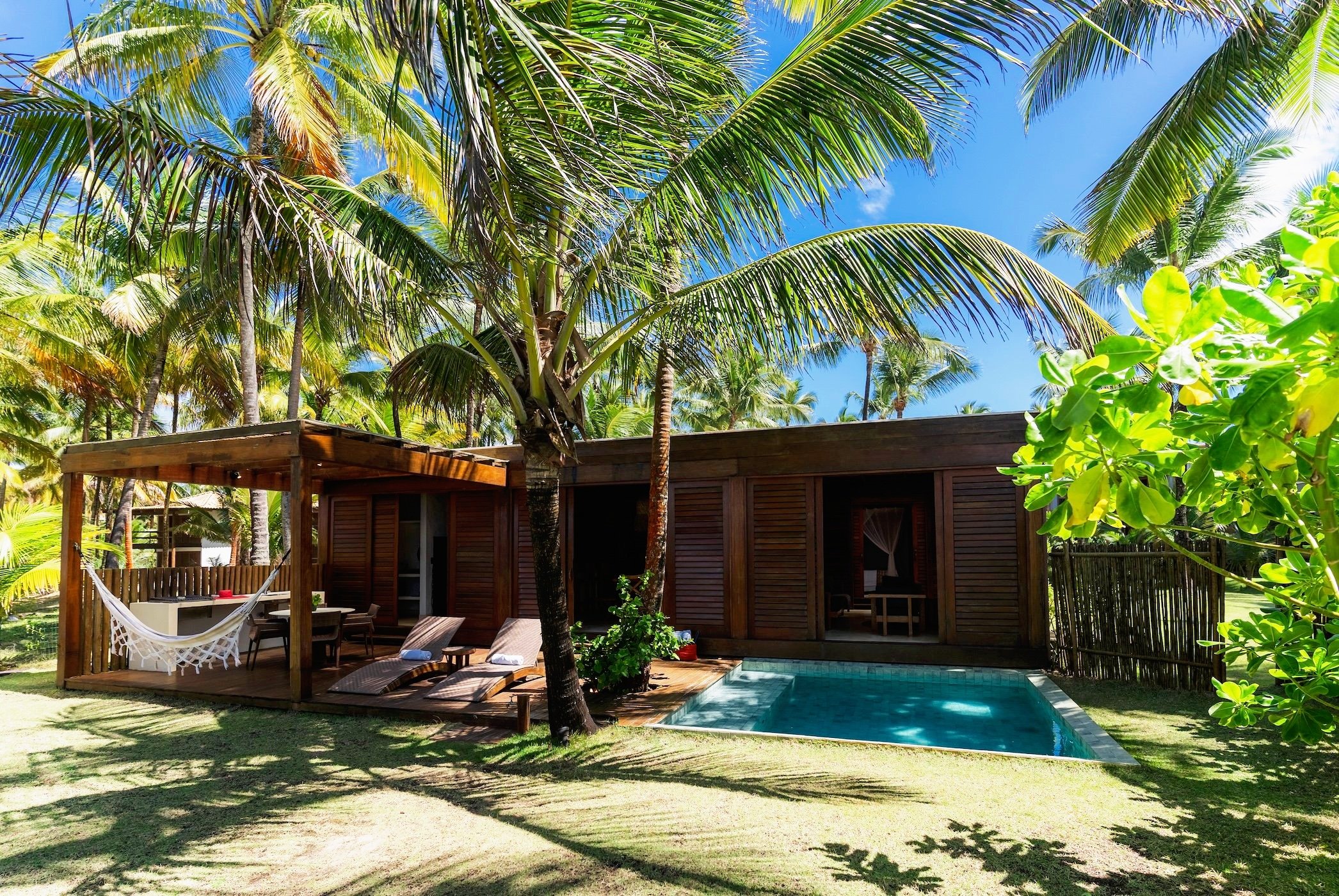 Casa de madeira com piscina e área de lazer ao ar livre sob guarda-sol, cercada por árvores e vegetação tropical, em clima ensolarado.