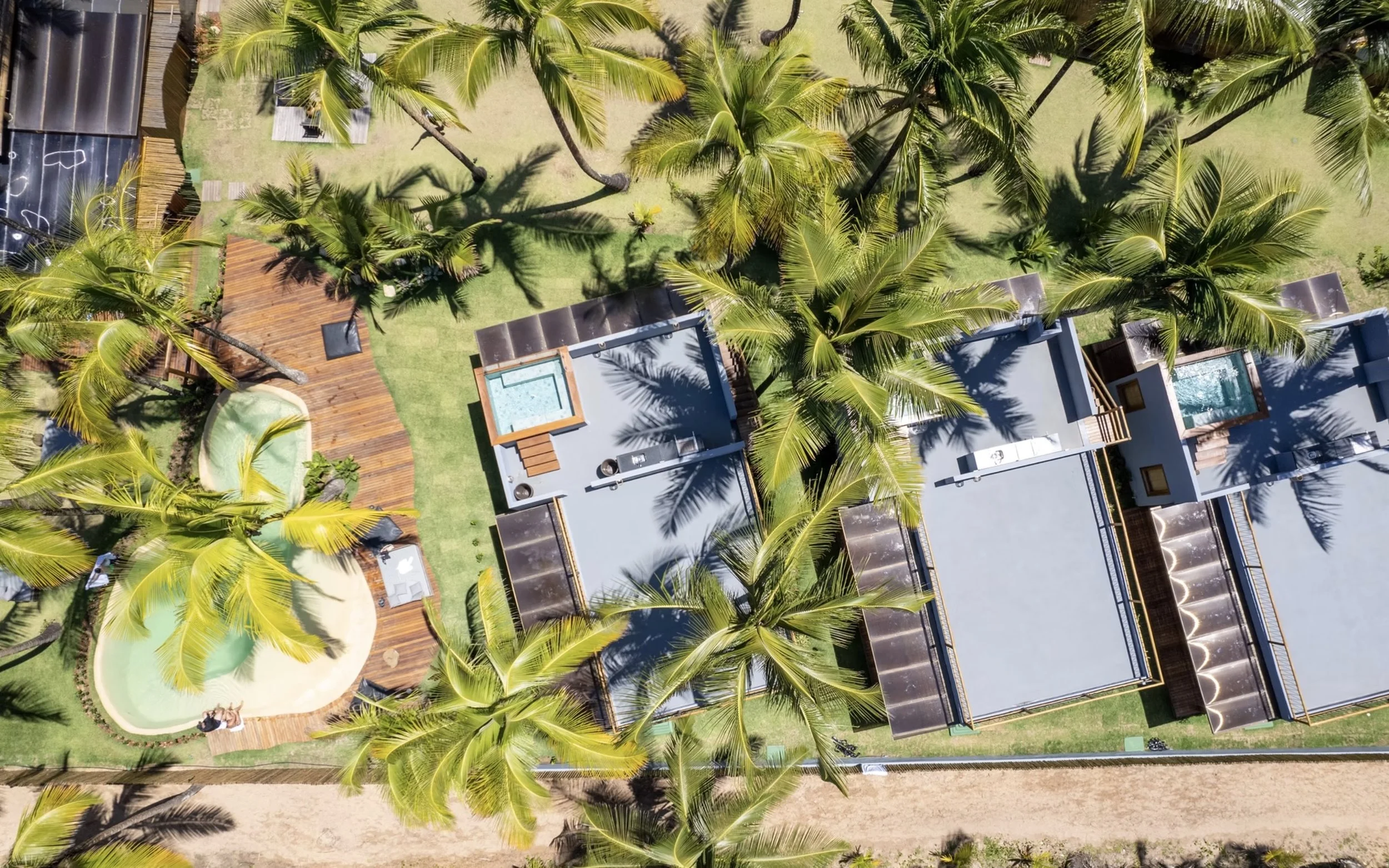 Imagem aérea de um condomínio de casas com telhados planos, cercado por muitas árvores de coqueiro e área de jardim, com duas piscinas visíveis.
