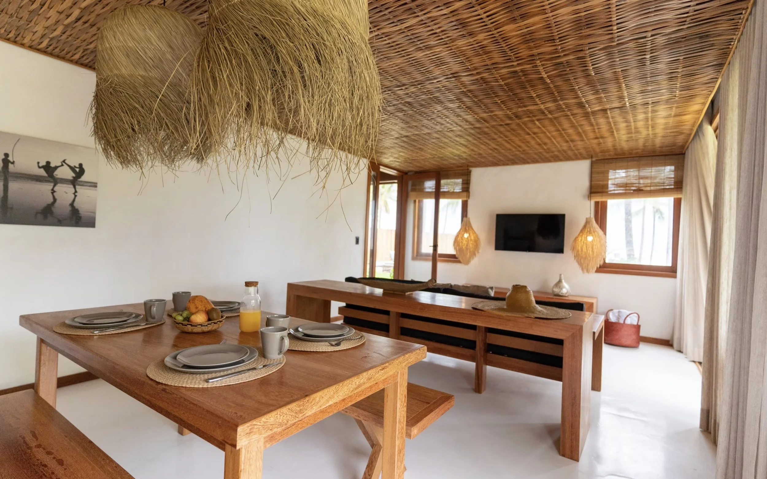 Sala de jantar decorada com móveis de madeira, luminárias de palha, mesa posta com pratos, copos e frutas, e televisão na parede