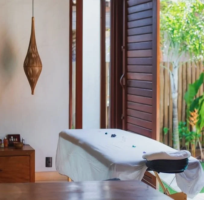 Sala de massagem com mesa coberta de toalha branca, pedras pequenas coloridas sobre a mesa, janela aberta com persianas de madeira ao lado, luminária suspensa de design moderno, e plantas do lado de fora.