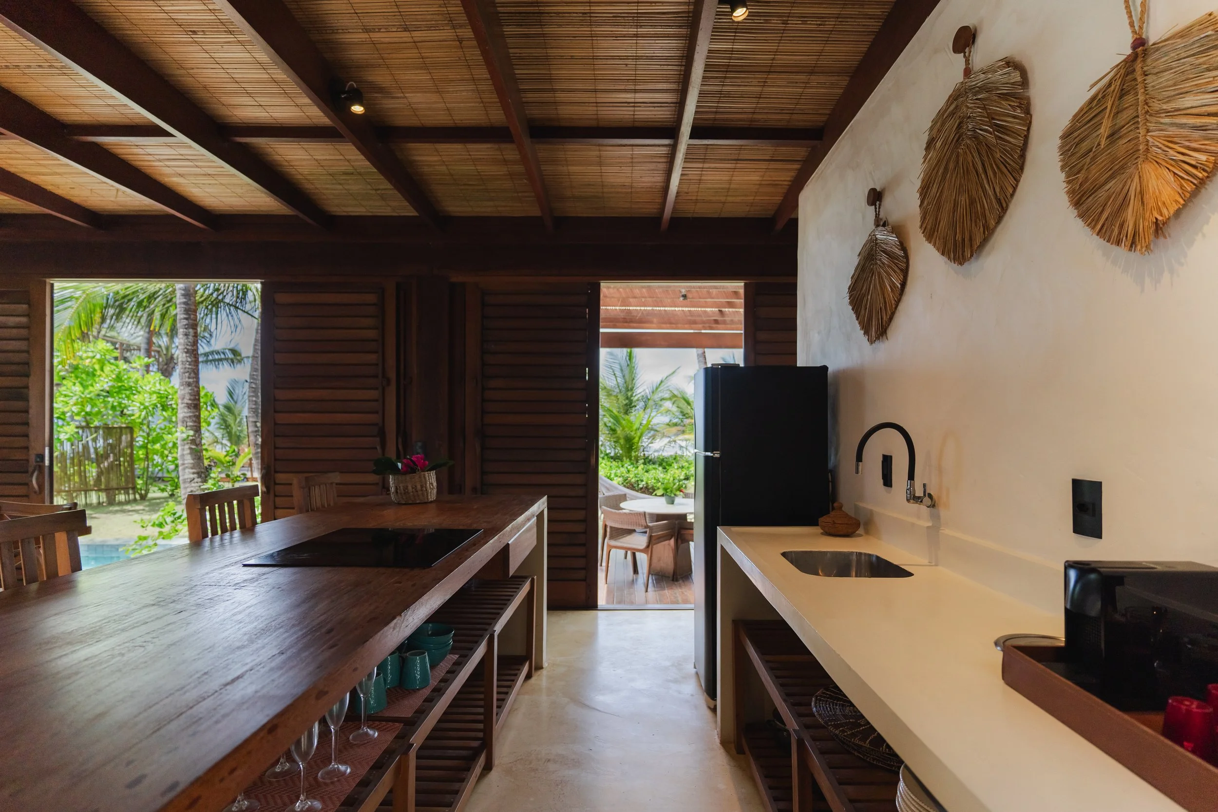 Interior de uma cozinha com bancada de madeira, parede de barro, decoração com folhas secas na parede, vista para um espaço externo com árvores, céu e piscina.