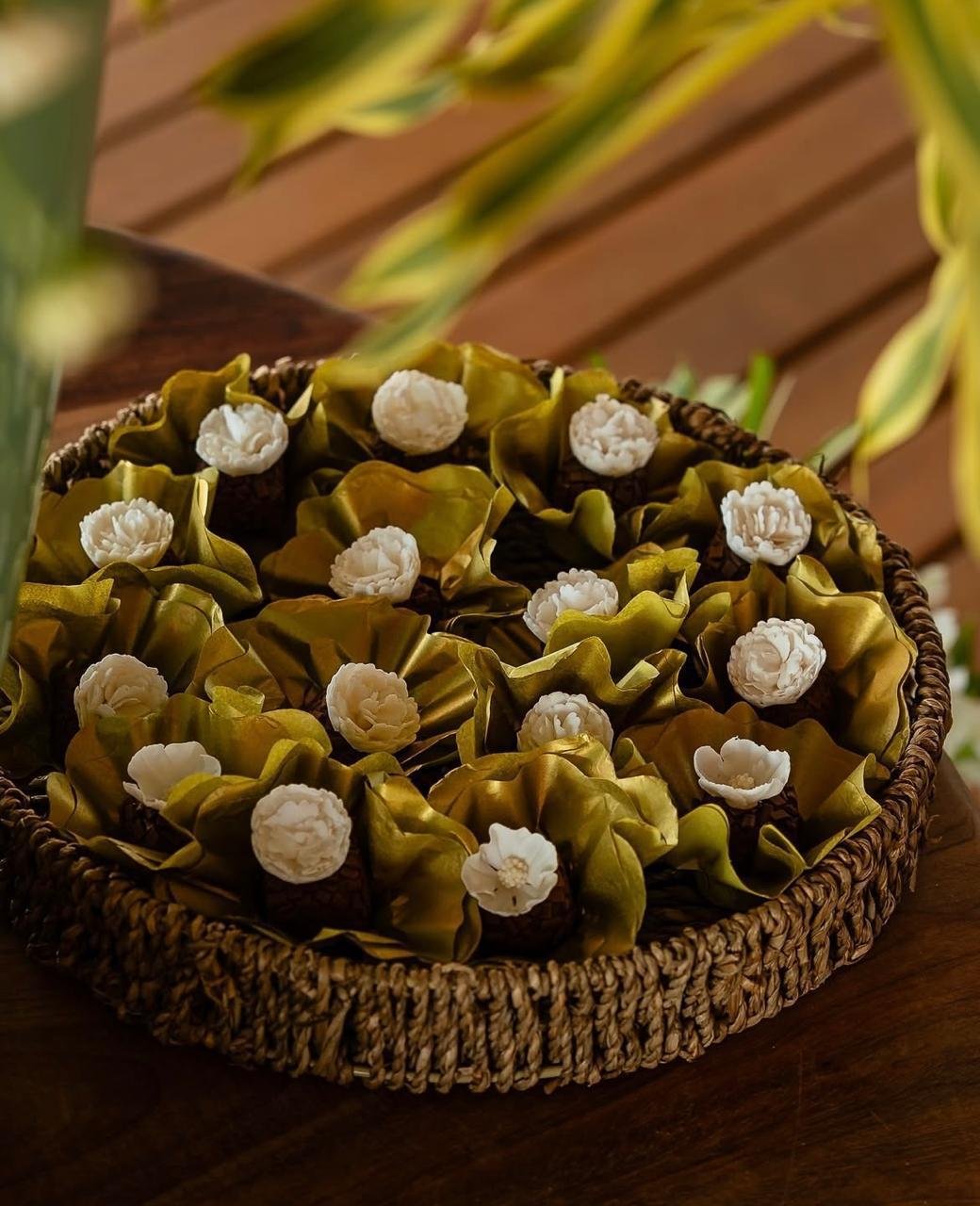 Vaso de bombons decorados com folhas verdes e flores brancas, colocado sobre uma superfície de madeira, rodeado por plantas verdes.
