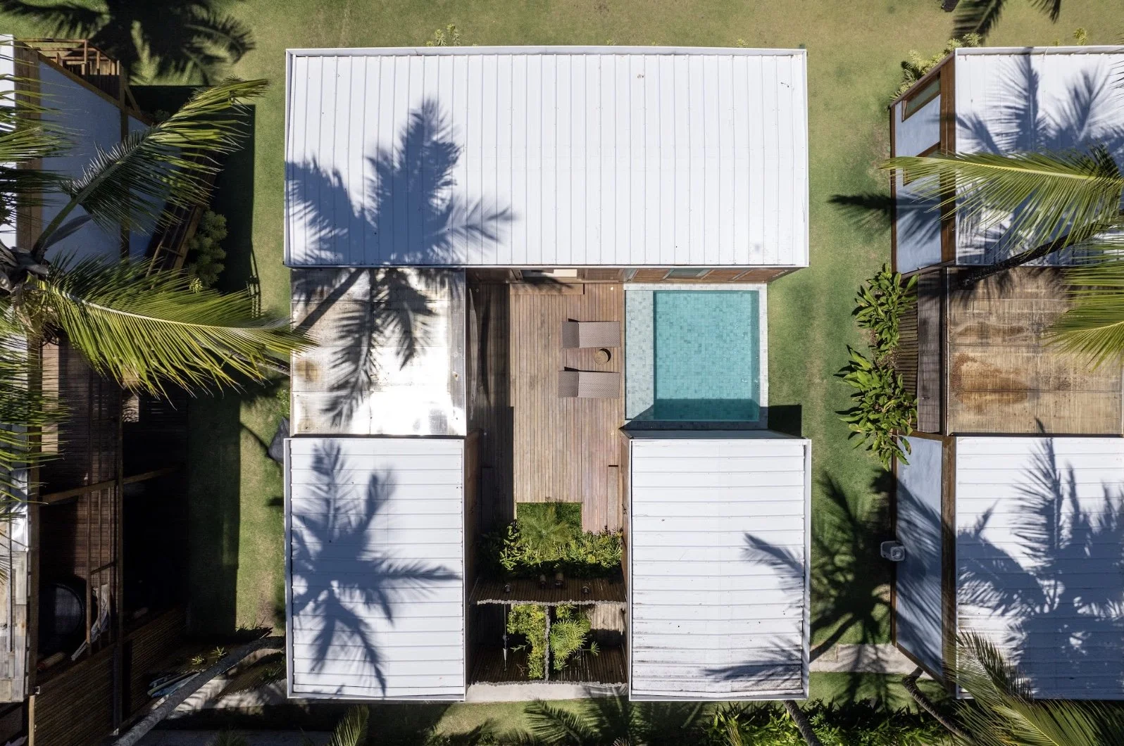 Vista aérea de casas com telhados brancos e piscina, cercadas por palmeiras e sombra das árvores, em área verde.