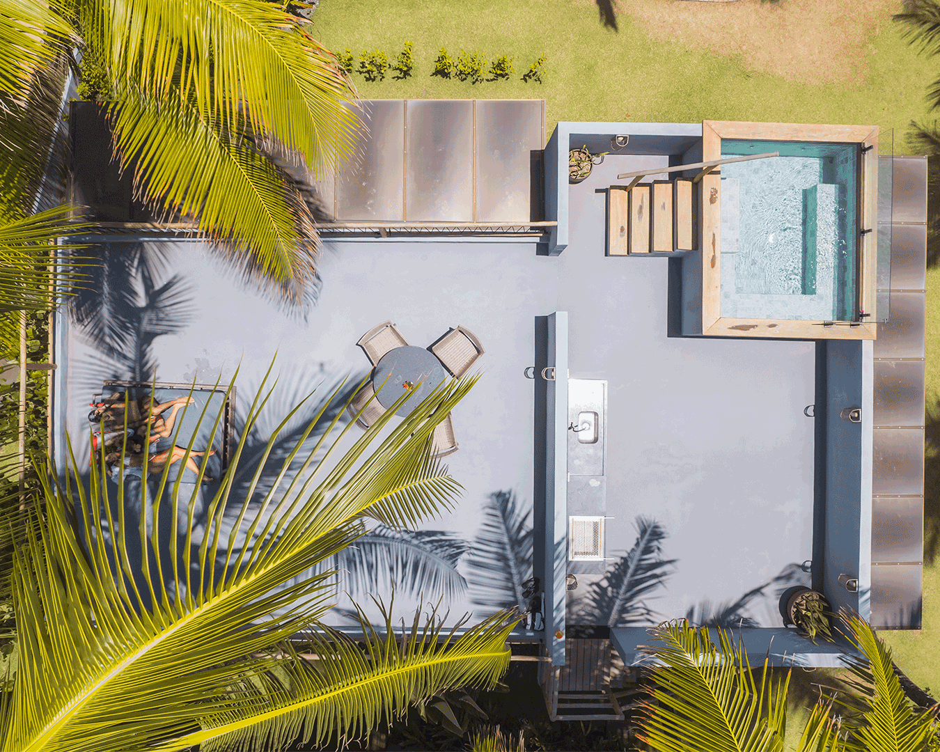 Vista aérea de uma área ao ar livre com uma piscina de borda rasa, uma mesa de jantar com cadeiras, plantas tropicais ao redor e um espaço coberto ao fundo, tudo em um ambiente de jardim bem cuidado.