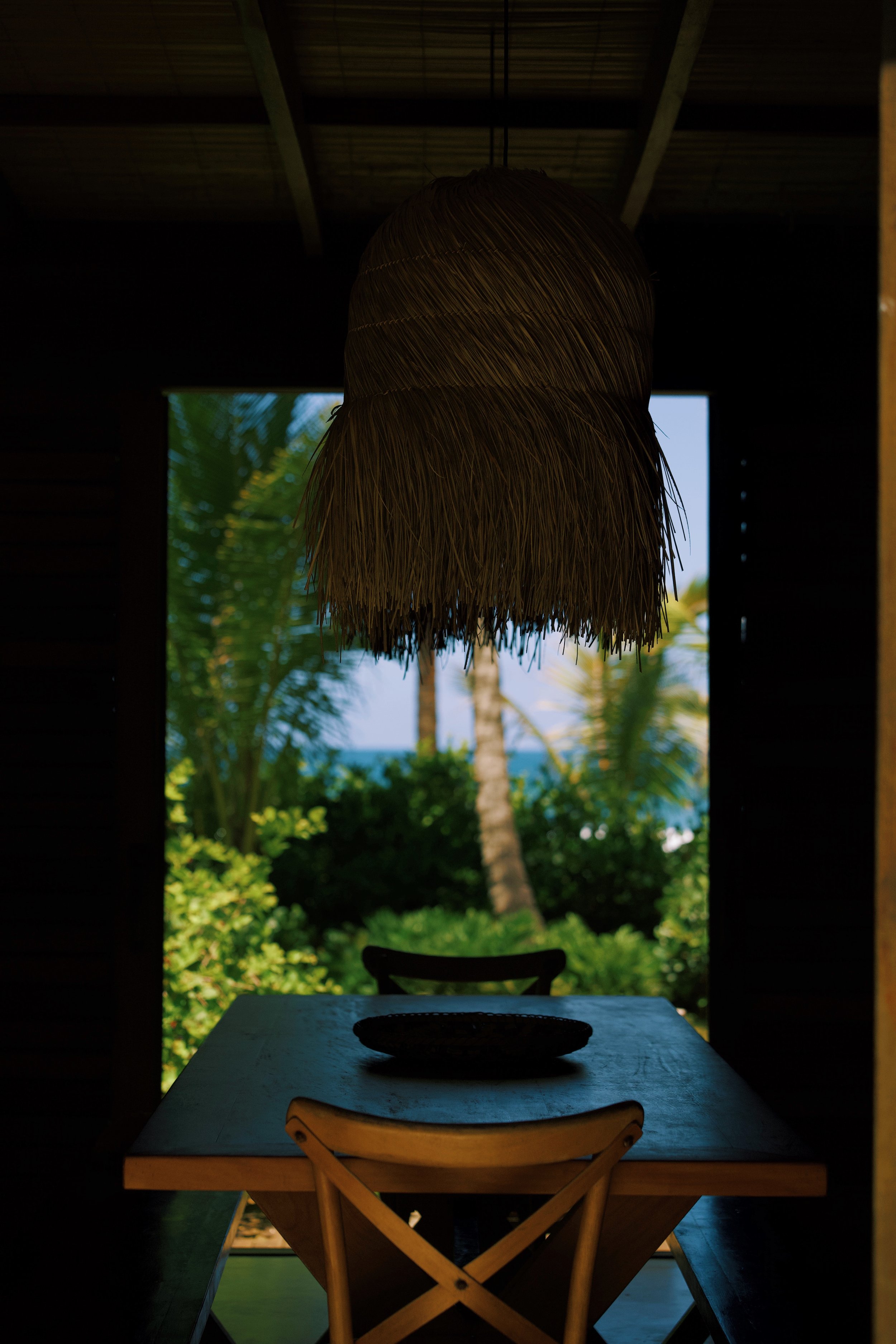 Cena do interior de uma cabana com uma mesa de madeira clara, uma cadeira de almoço e um prato sobre ela, na frente de uma janela que revela uma paisagem tropical com palmeiras, árvores verdes e o mar ao fundo.