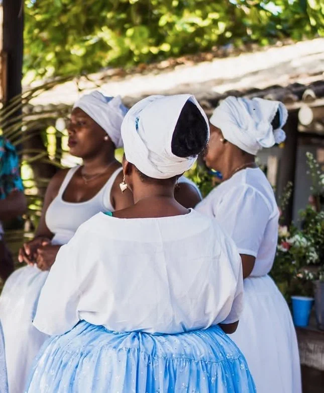 Grupo de mulheres vestidas com roupas tradicionais brancas e turbantes brancos em ambiente externo com vegetação verde ao fundo.