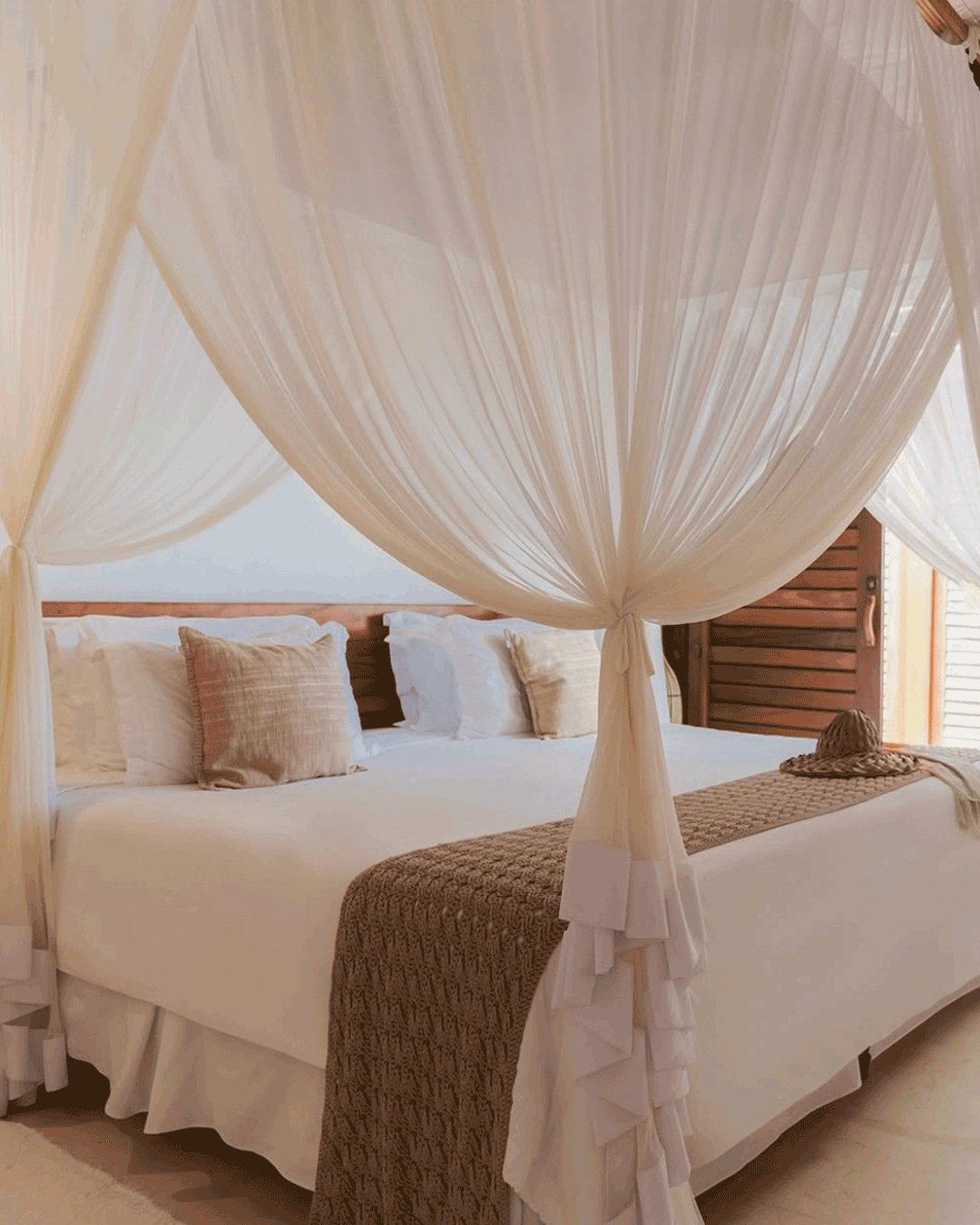 Quarto com cama com dossel de tule branco, travesseiros bege e branco, cobertor castanho na cama, e janela com persianas de madeira ao fundo.