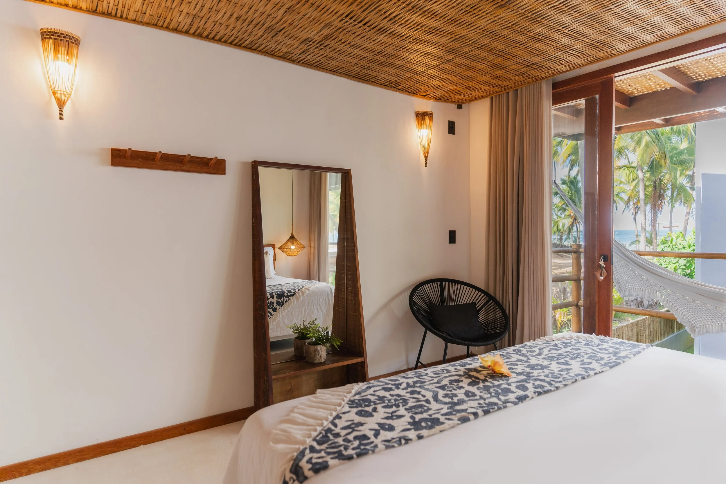Quarto de decoração tropical com teto de palha, paredes brancas, iluminação de parede em estilo cabaña, espelho de madeira, planta em vaso ao lado da cama, cadeiras de design moderno, vista para árvores e praia através de uma grande janela.