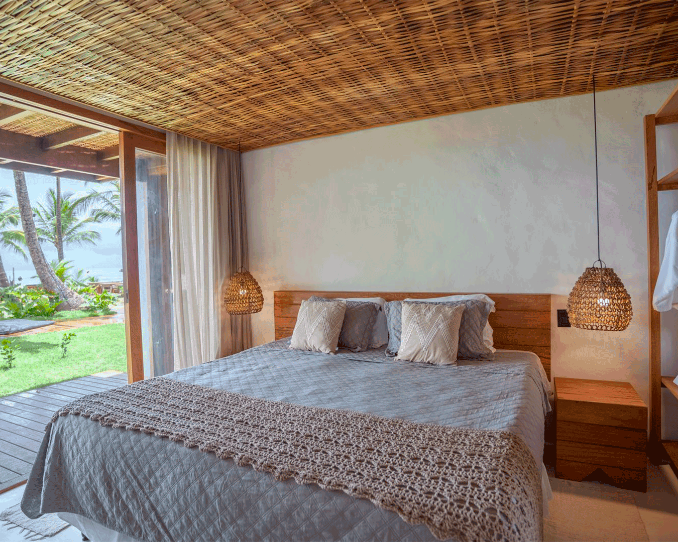 Quarto com cama de madeira, roupa de cama cinza, quatro travesseiros, luminárias de balanço, vista de uma varanda com plantas tropicais e árvores ao ar livre.