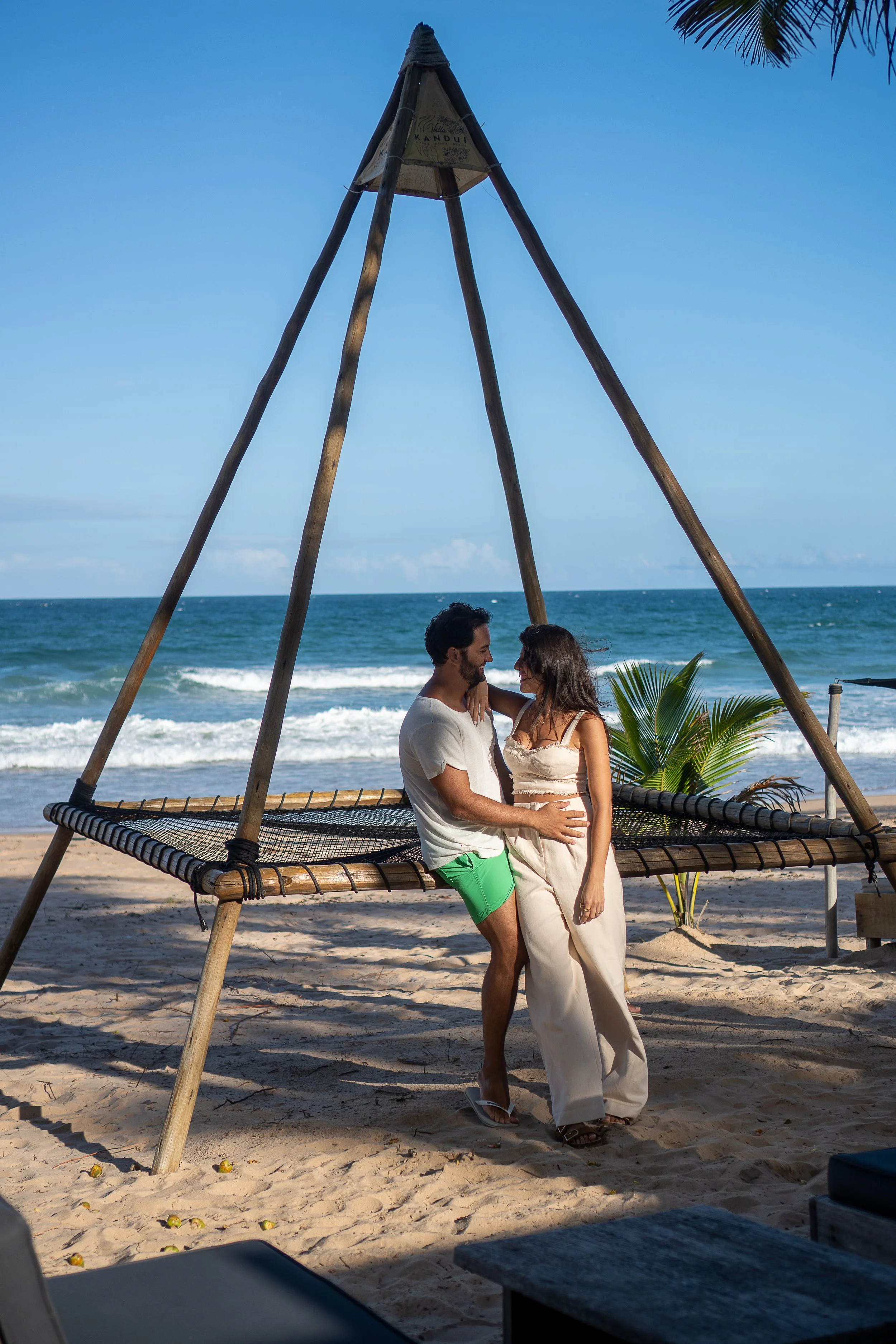 Casal no litoral, de frente, sorrindo, sob céu azul, com windsurf de madeira na areia e mar ao fundo, criando uma atmosfera romântica na praia.