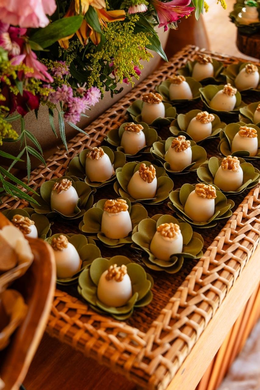 Docinhos decorados com castanhas, servidos em pequenas folhas verdes, dispostos em uma bandeja de palha, ao lado de arranjo de flores coloridas.
