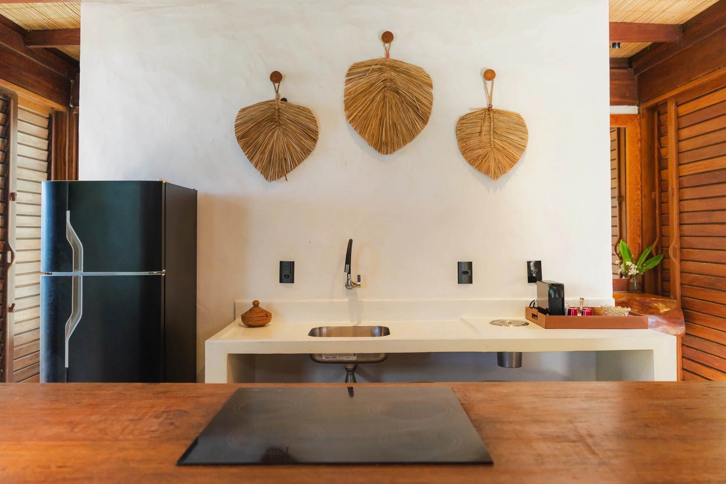 Parede branca com três, folhas decorativas de palha suspensas, ao lado de uma bancada com centro de pia, utensílios e uma geladeira preta, com portas de madeira ao redor.