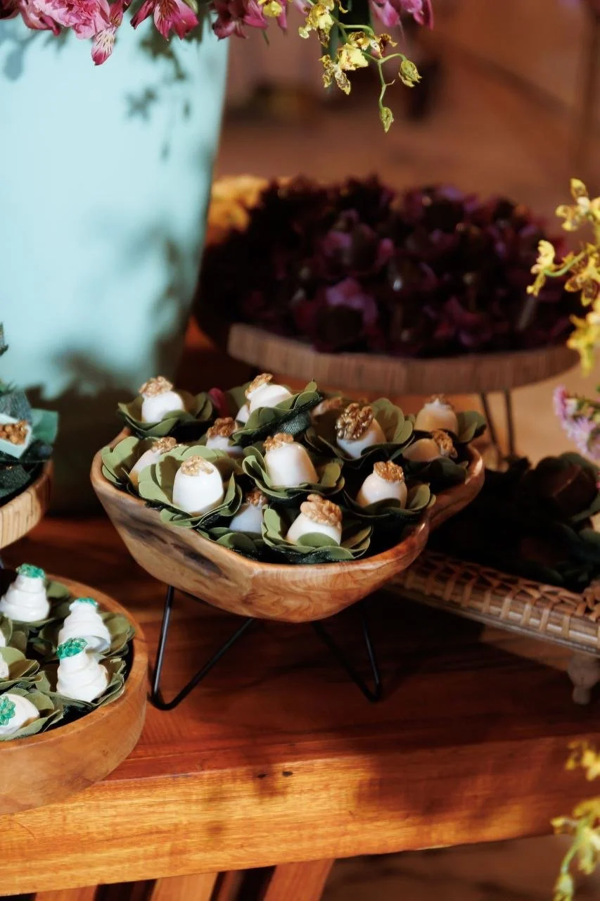 Diversas sobremesas em taças de vidro, decoradas com folhas verdes e castanhas, dispostas em travessas de madeira na mesa.