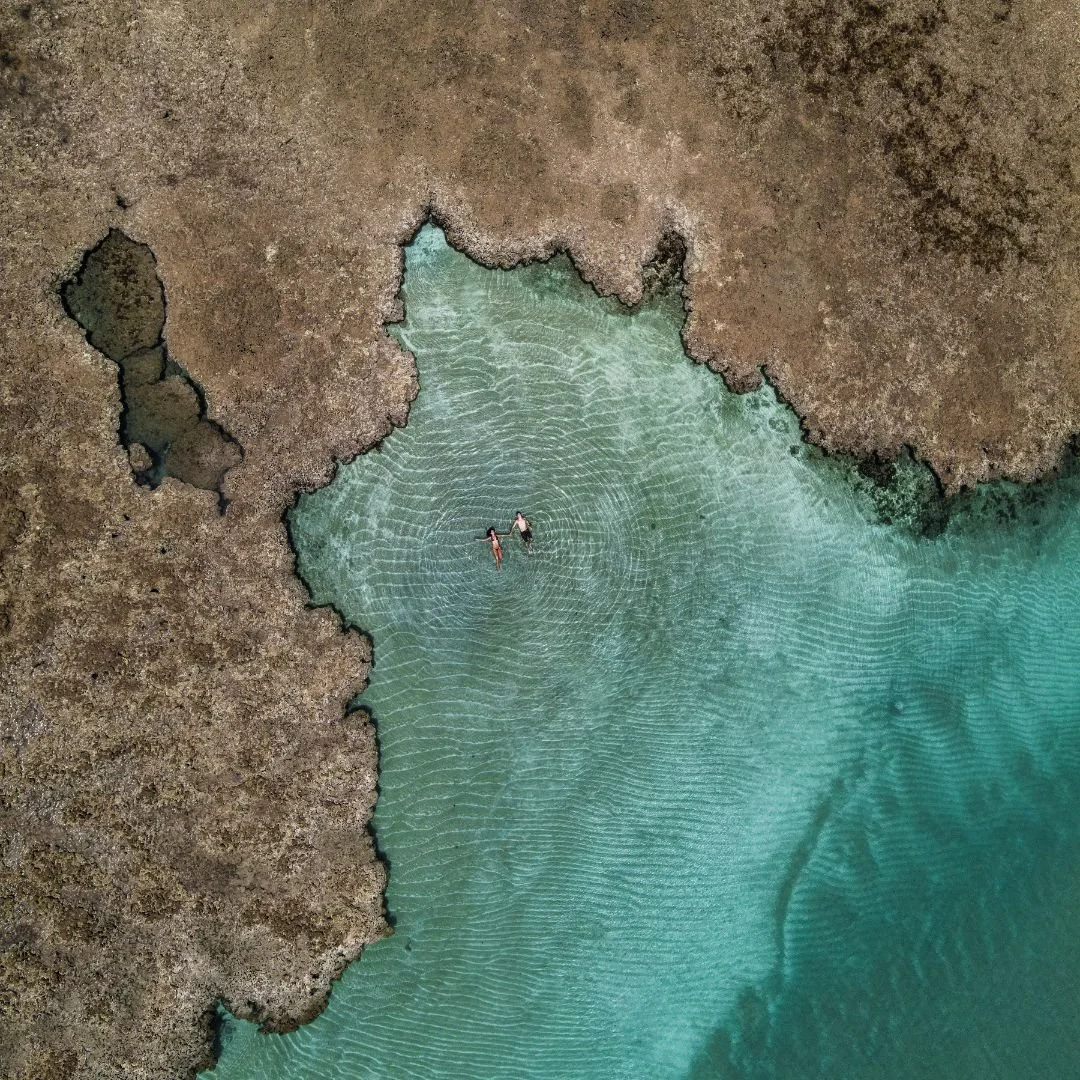 Vista aérea de duas pessoas nadando em uma piscina natural cercada por rochas e água clara. A imagem mostra um contraste entre o terreno rochoso e a água azul-turquesa.