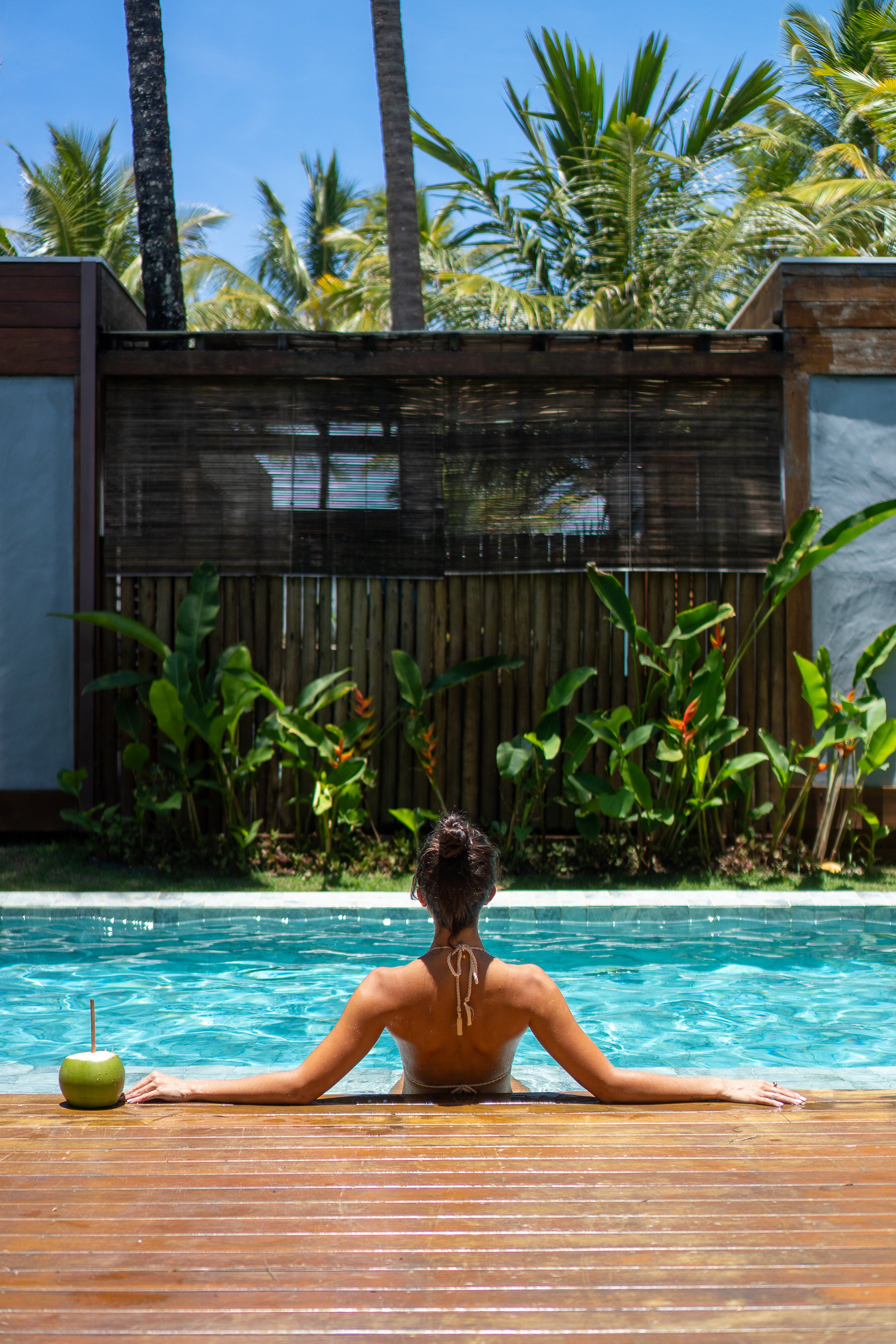 Mulher relaxando na piscina ao lado de uma Coco gelada, com vegetação tropical ao fundo e céu azul.