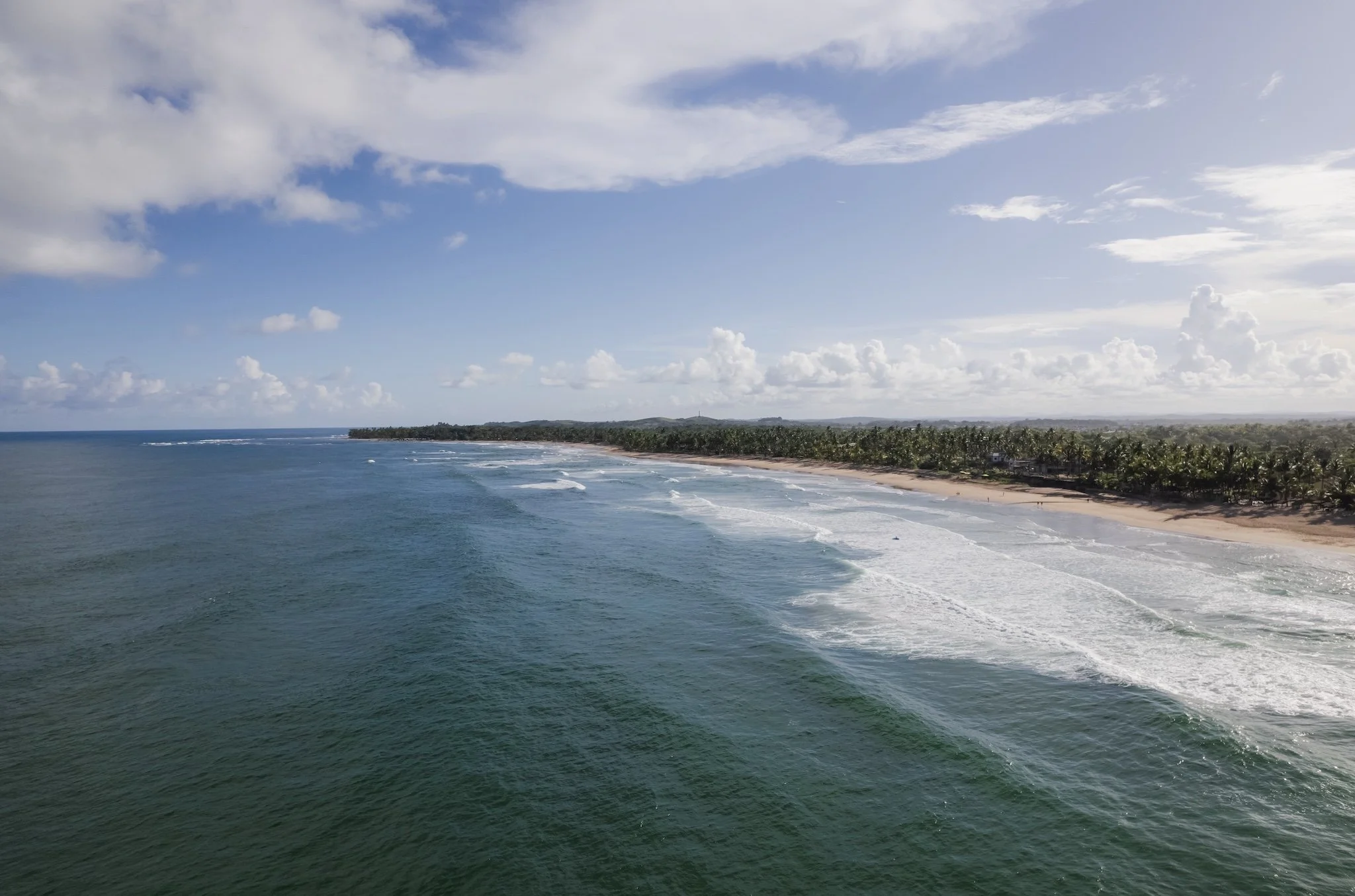 Praia com mar azul e ondas, céu azul com nuvens, faixa de areia e vegetação ao fundo.