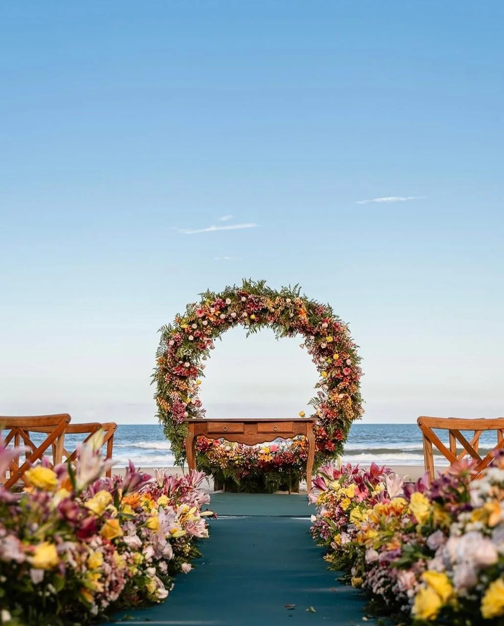 Caminho de casamento decorado com flores, levando a uma estrutura de arco de flores na praia com o mar ao fundo.