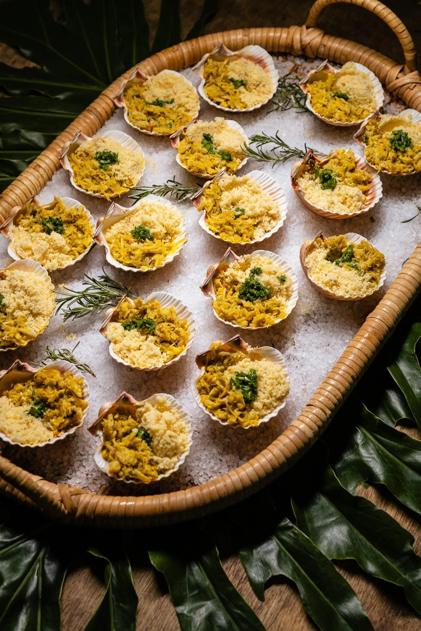 Cubos de marisco com queijo, decorados com alecrim, servidos em conchas, dispostos sobre sal grosso em uma bandeja de palha, com folhas verdes ao redor.