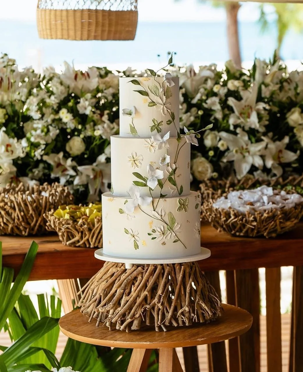 Bolo de casamento de três andares branco decorado com flores brancas e galhos verdes, colocado sobre uma base de troncos de madeira, em uma mesa de madeira, com fundo de flores brancas e uma área ao ar livre.