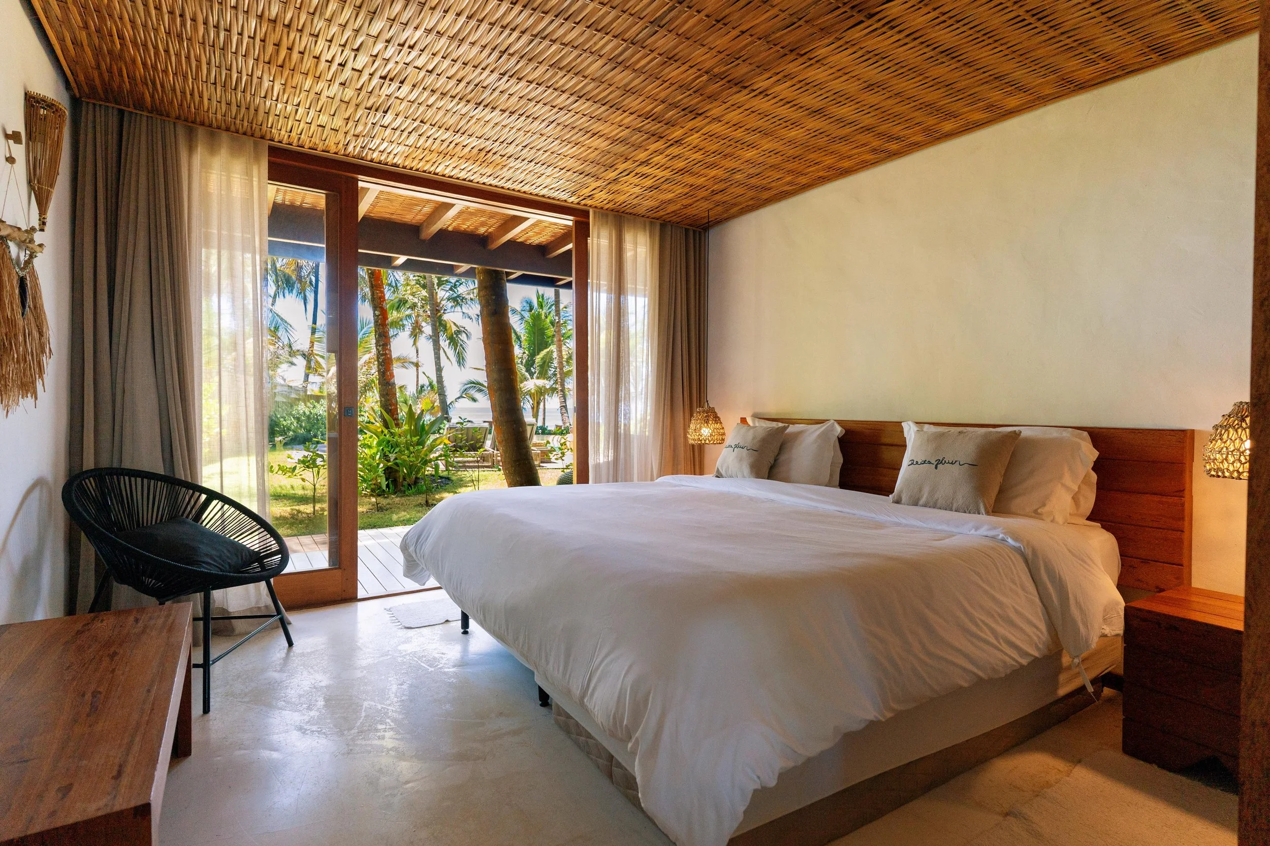 Quarto de hotel com cama de casal, travesseiros, luminária de cabeceira, parede de pedra, cortinas abertas mostrando varanda com vista para árvores tropicais.