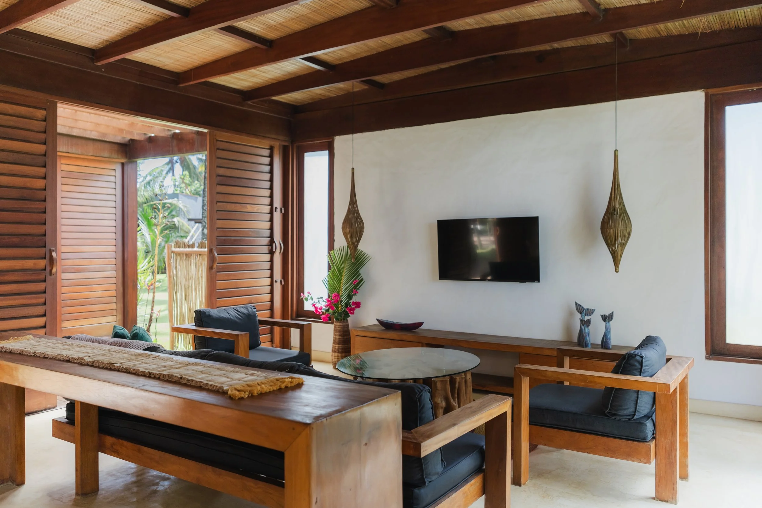 Sala de estar com móveis de madeira, poltrona de couro preta, mesa de centro de vidro, plantas e janela com cortinas de madeira, decorada com vasos e objetos de decoração.