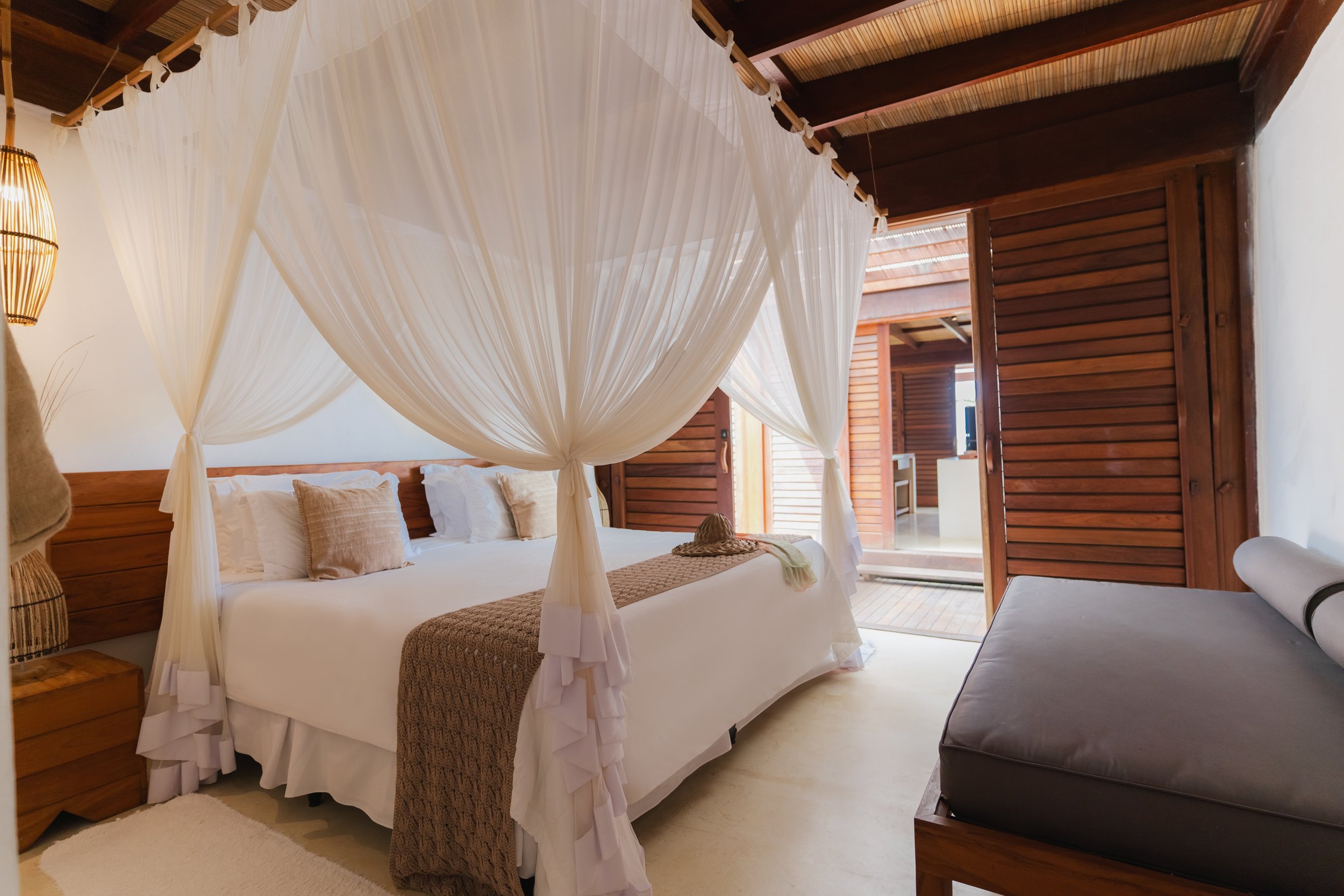Quarto de hotel com cama de casal, cortina de tecido branco decorativa na cama, móveis de madeira, papel de parede de madeira, parede branca, luminária de pêndulo de palha e uma porta de correr de madeira.