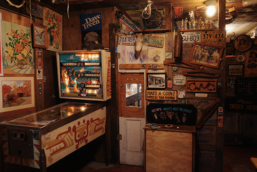 Vintage pinball machine in a cozy, rustic bar or cafe decorated with various paintings, license plates, and memorabilia, including a mounted skull and framed photographs.