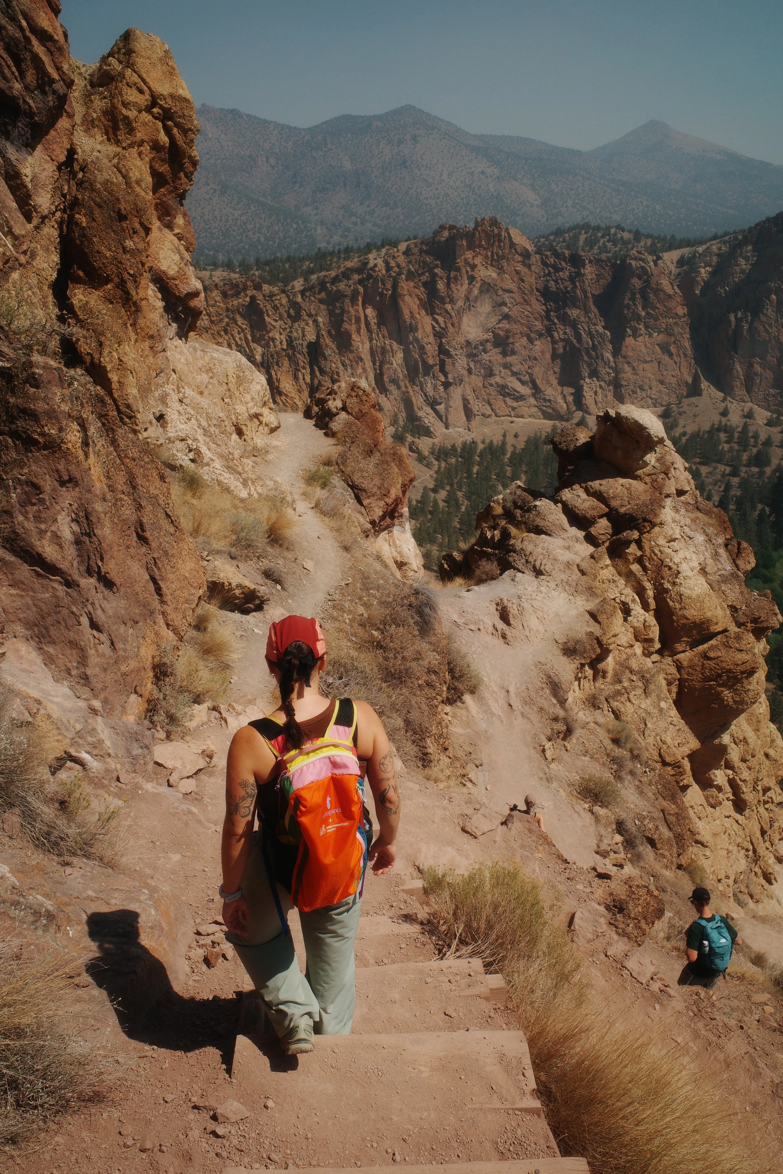 Hikers walking along a rocky mountain trail with a desert landscape and mountains in the background.
