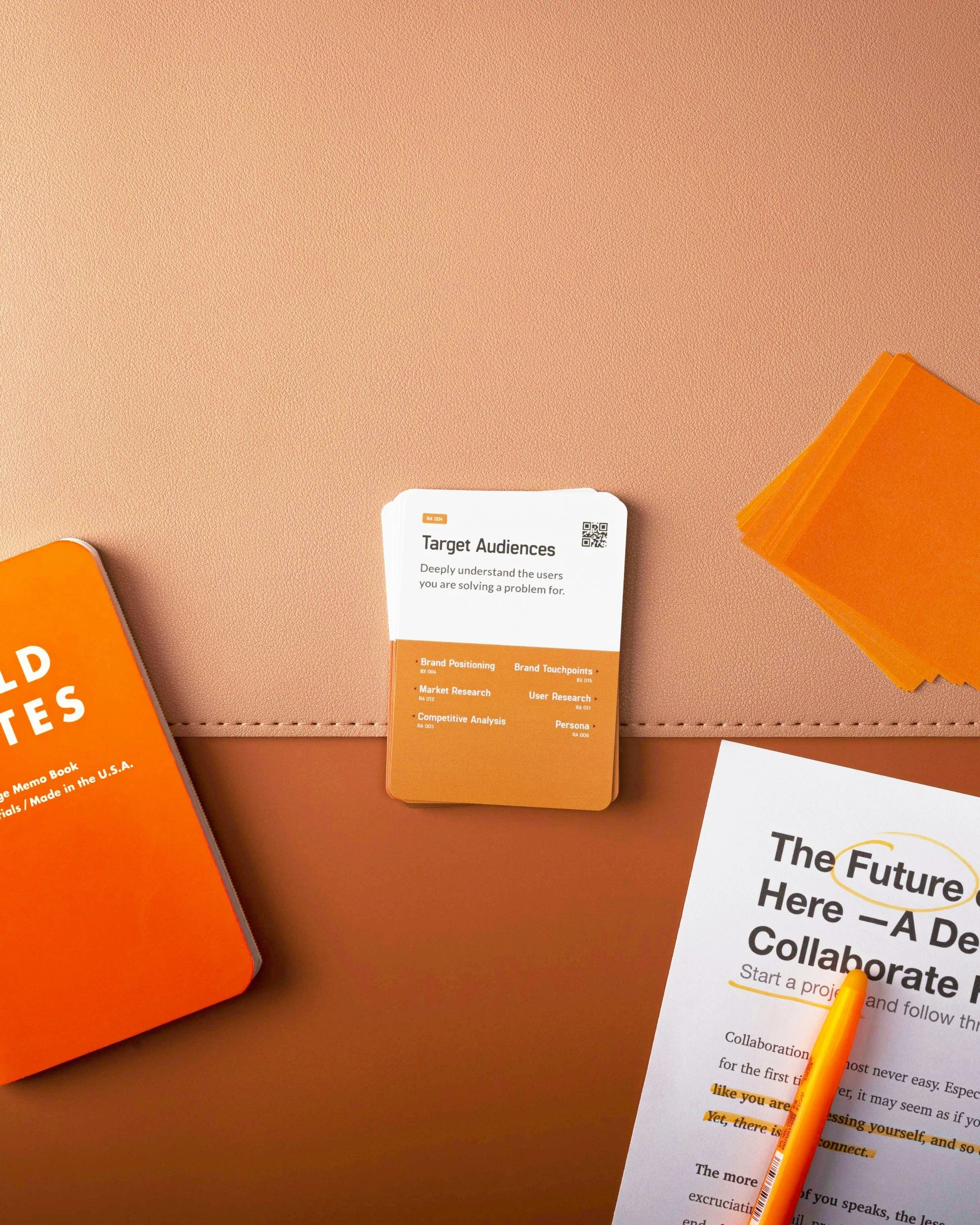 A conference or presentation table with orange and white materials, including a card titled 'Target Audiences' with bullet points on market research and brand positioning, an orange notebook, and a white paper with text about collaboration and project start, along with a yellow pen.