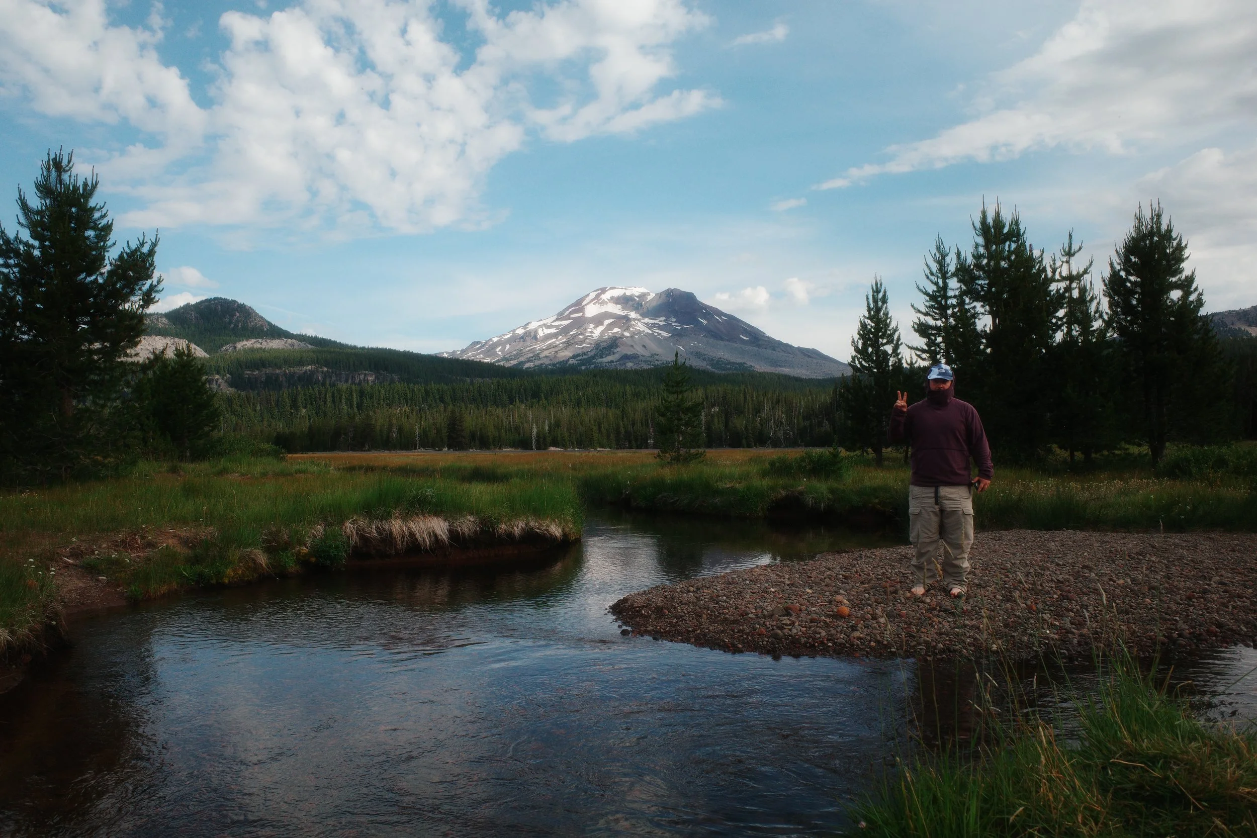 Person standing on rocky bank by a calm river in a forested area with snow-capped mountain in the background, partially cloudy sky.