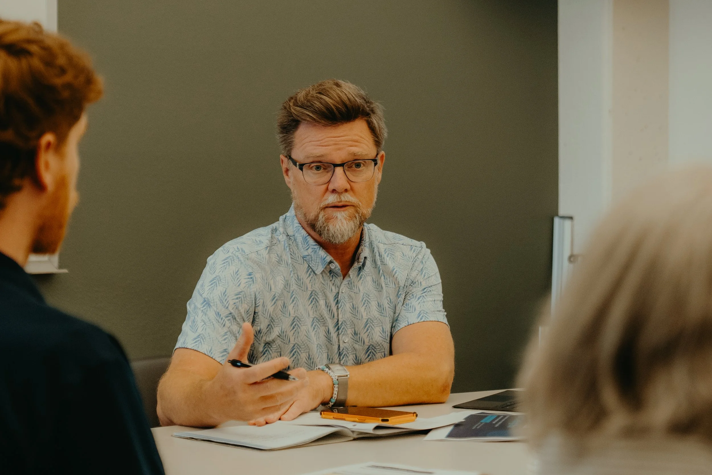 A middle-aged man with glasses, a beard, and mustache wearing a light patterned shirt, sitting at a table during a discussion, with papers, a smartphone, and a laptop in front of him. He is engaging with two others, partly visible in the foreground, in an indoor setting with a gray wall background.