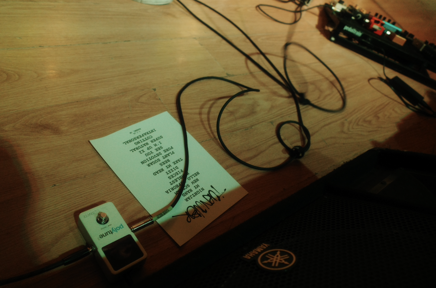 Close-up of a wooden table with a small portable device connected to a string, a printed setlist, and a partially visible Yamaha speaker in the bottom right corner.