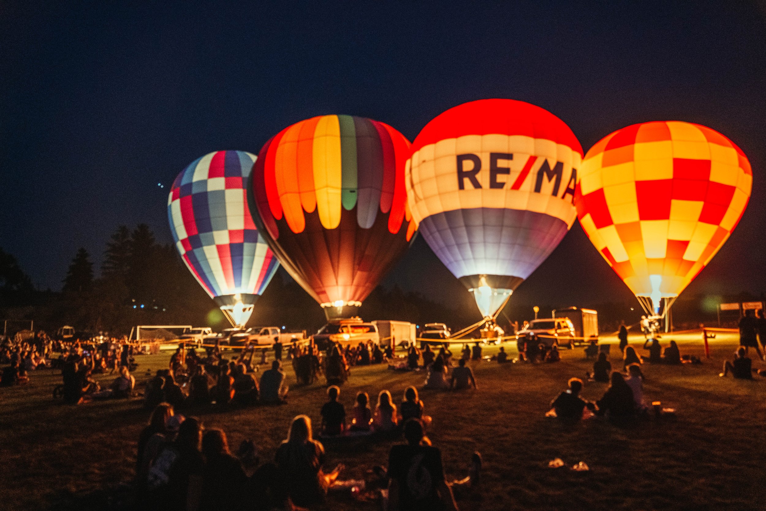 Nighttime scene of hot air balloons being inflated and prepared for ascent at a festival, with people sitting and standing on the grass and cars parked nearby.