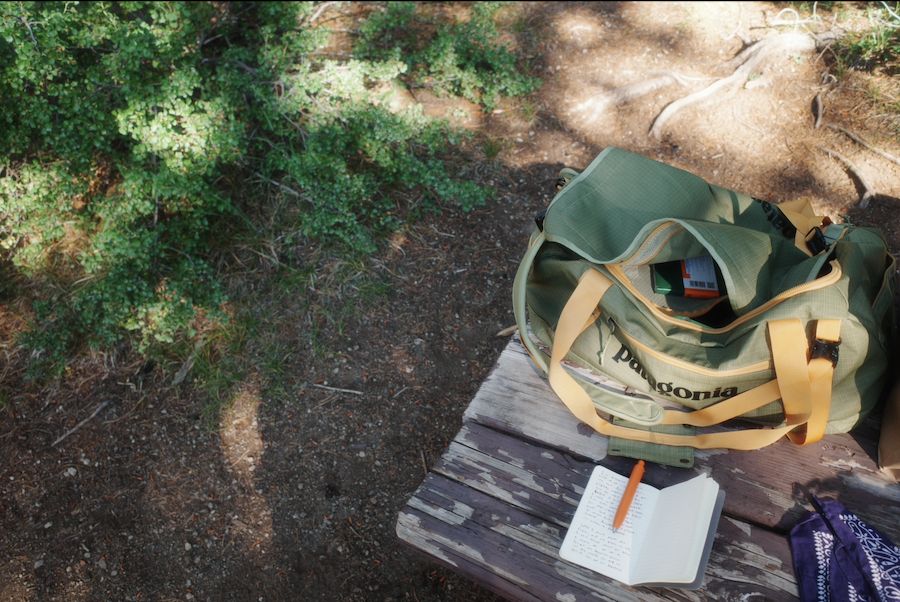 A green and yellow Patagonia duffel bag on a weathered wooden bench in a natural outdoor setting. The bag is open, revealing some books inside. Next to the bag, there is a small notebook with handwritten notes and an orange pen on top. The ground is dirt, with a nearby shrub casting a shadow.