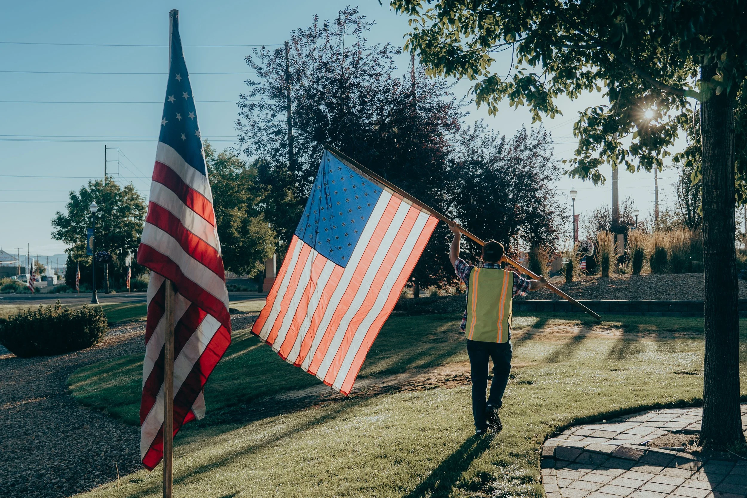 Person in a yellow vest holding a large American flag on a pole outdoors, with additional American flags on flagpoles nearby, trees, and a road in the background.