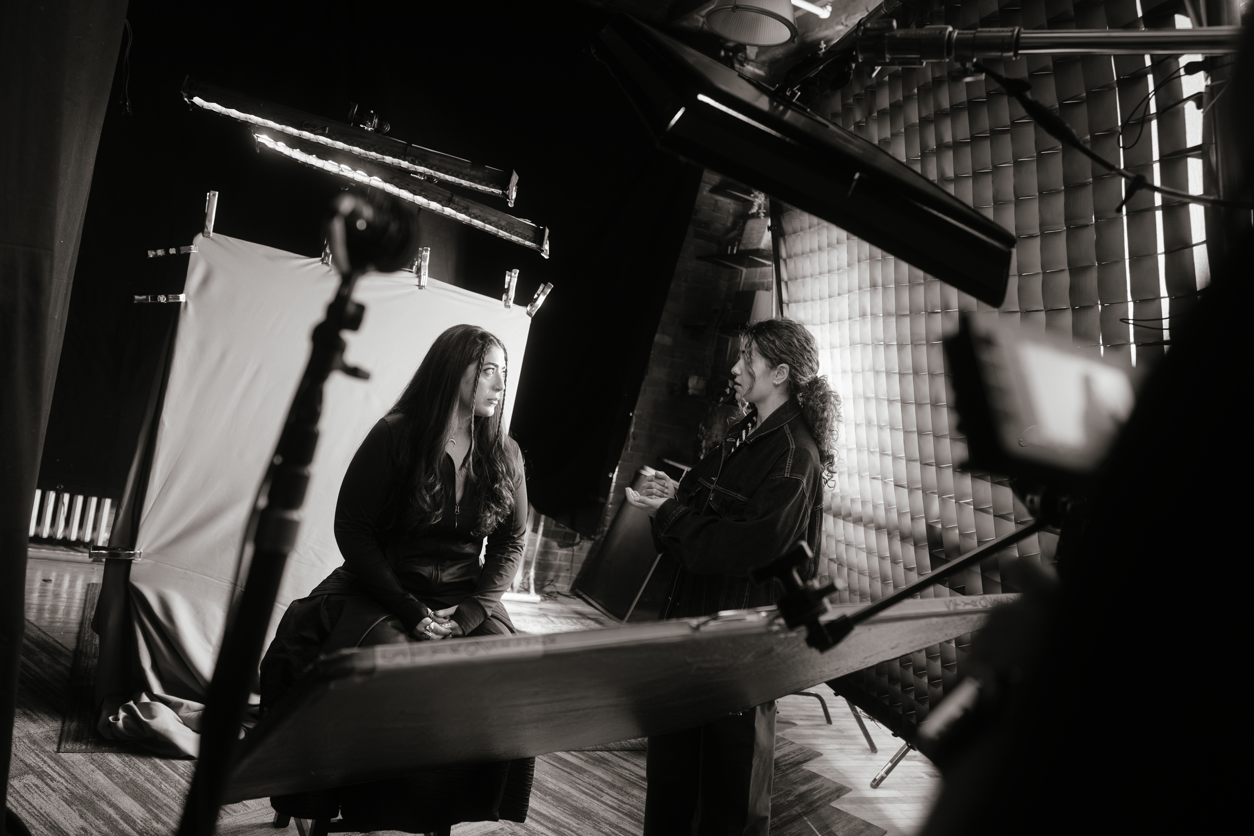 Black and white photo of a woman sitting on the floor in front of a plain backdrop, being interviewed by another woman in a studio setting with studio lights and equipment visible.