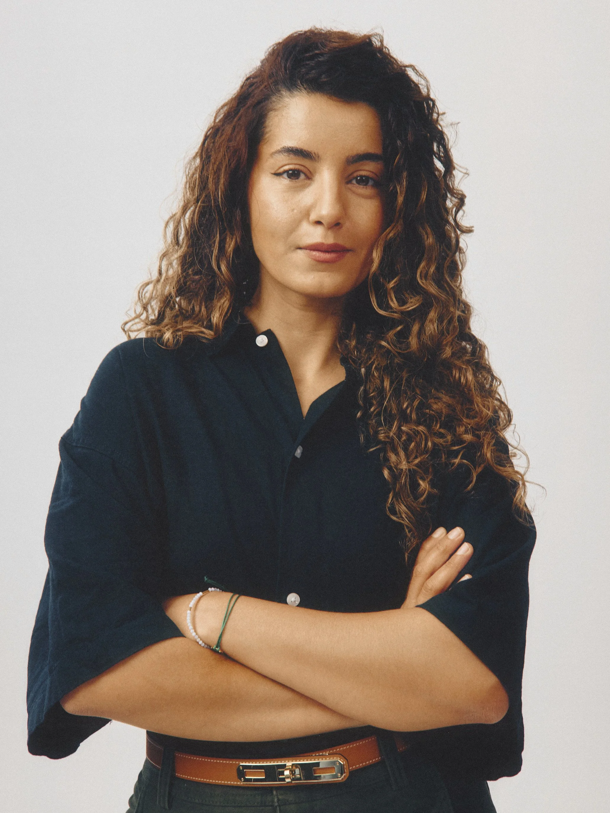 A woman with long, curly brown hair wearing a black jacket with rolled sleeves, a brown belt, and bracelets on her wrist, standing with arms crossed against a plain white background.