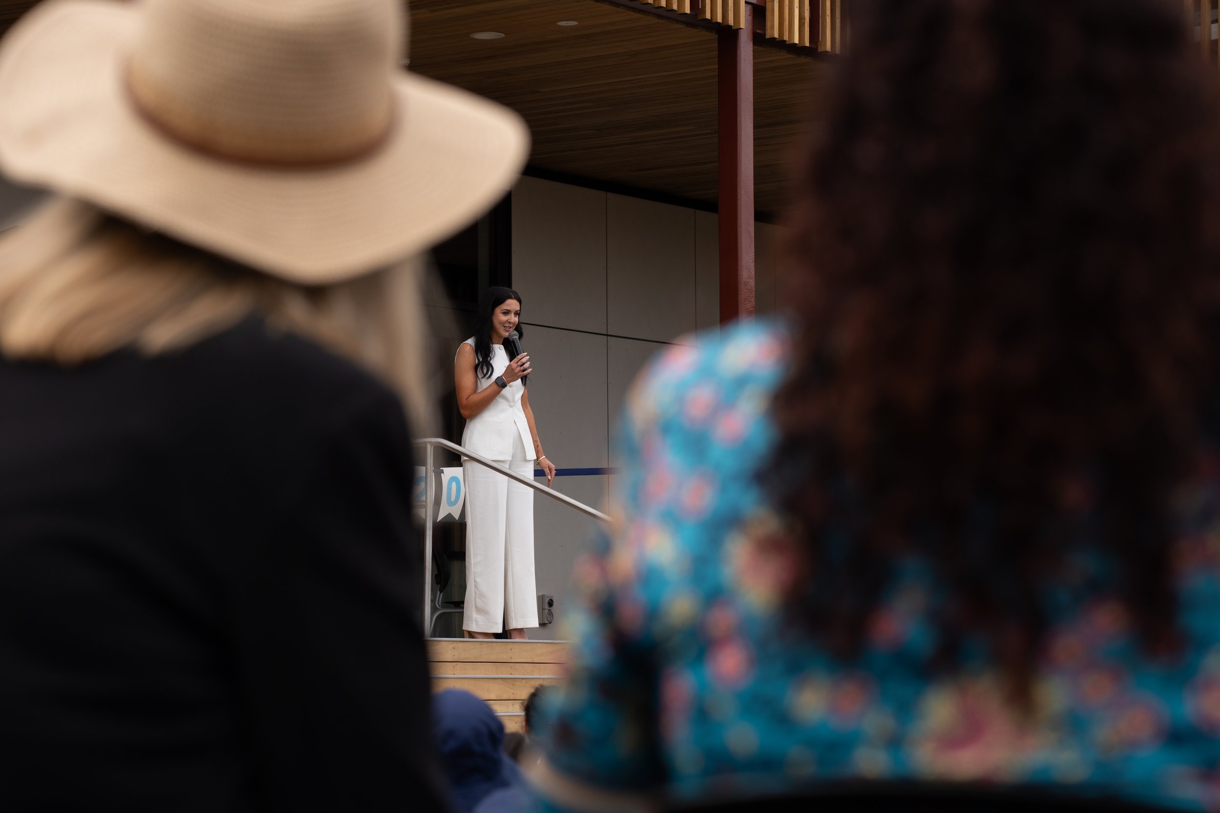 A woman in white pants and a sleeveless top speaking into a microphone on stage, seen through the heads of an audience, including a person wearing a wide-brimmed hat and another in a floral patterned shirt.