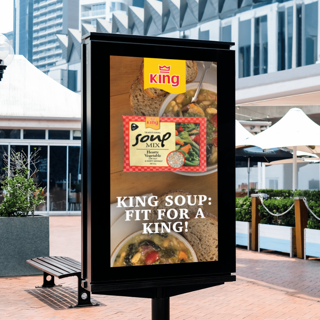 An outdoor advertisement board displaying a promotional poster for King Soup, featuring a bowl of vegetable soup, slices of bread, and text that says ''King Soup: Fit for a King!''