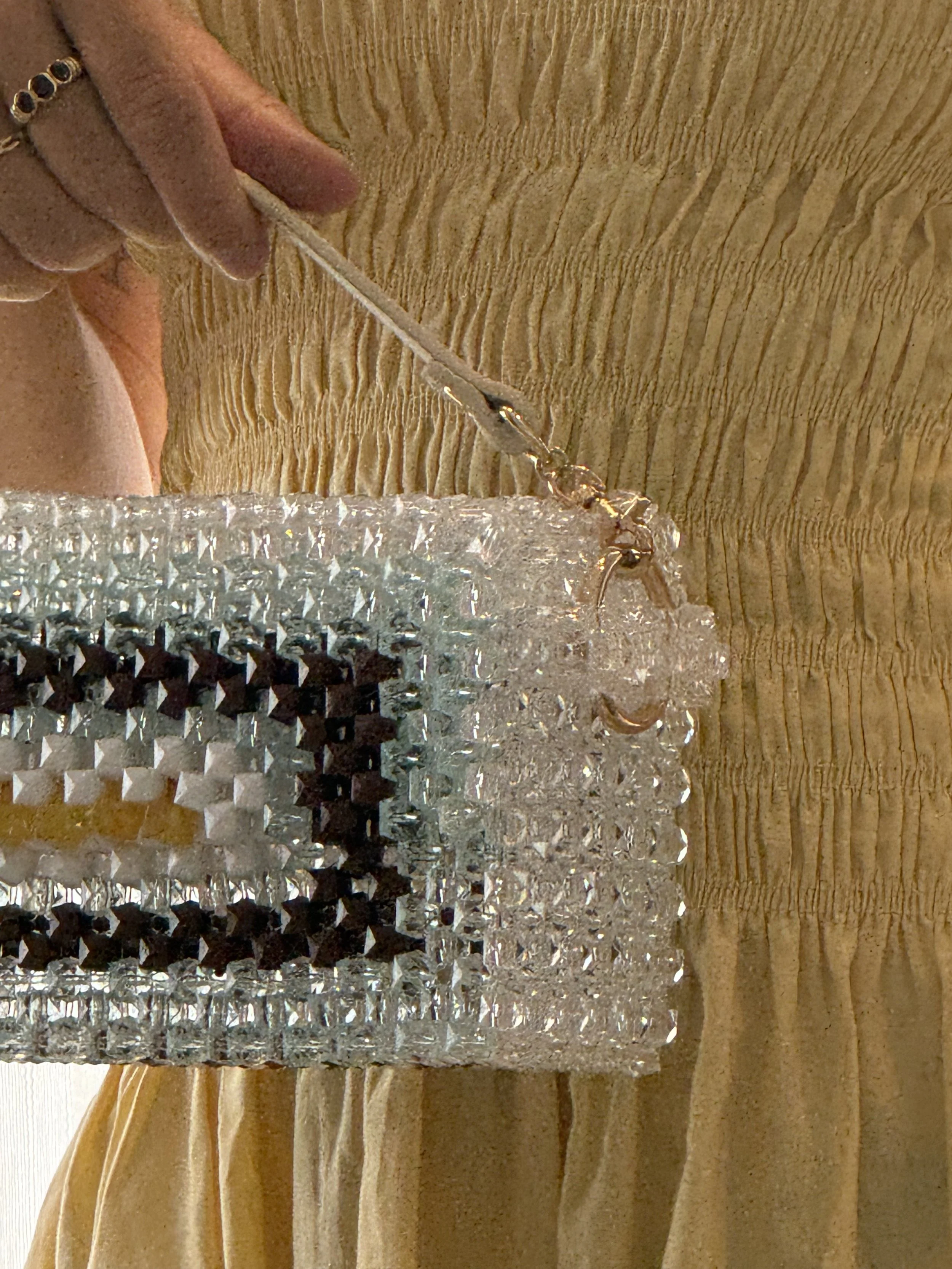 Close-up of a hand holding a silver chain link strap attached to a rectangular, textured, metallic silver clutch purse with black and clear rhinestones, against a beige, ribbed fabric background.