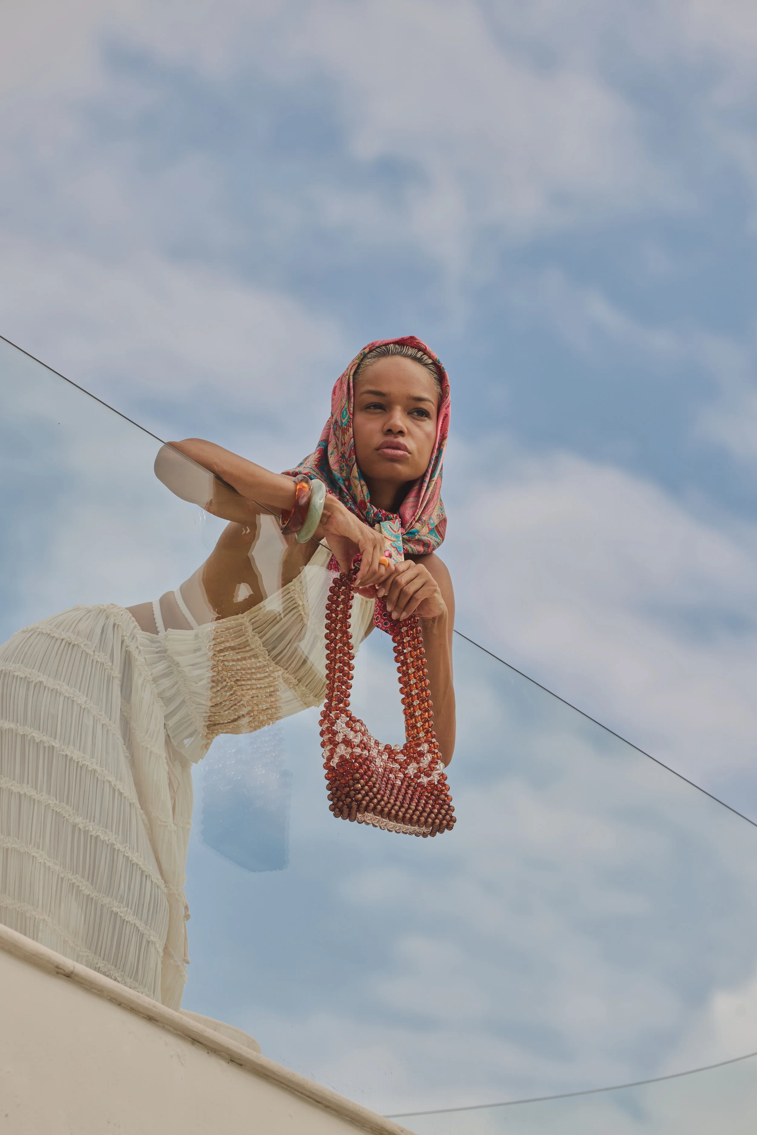 A woman wearing a colorful headscarf and a cream-colored dress, holding a beaded bag, leaning over a ledge with a cloudy sky in the background.