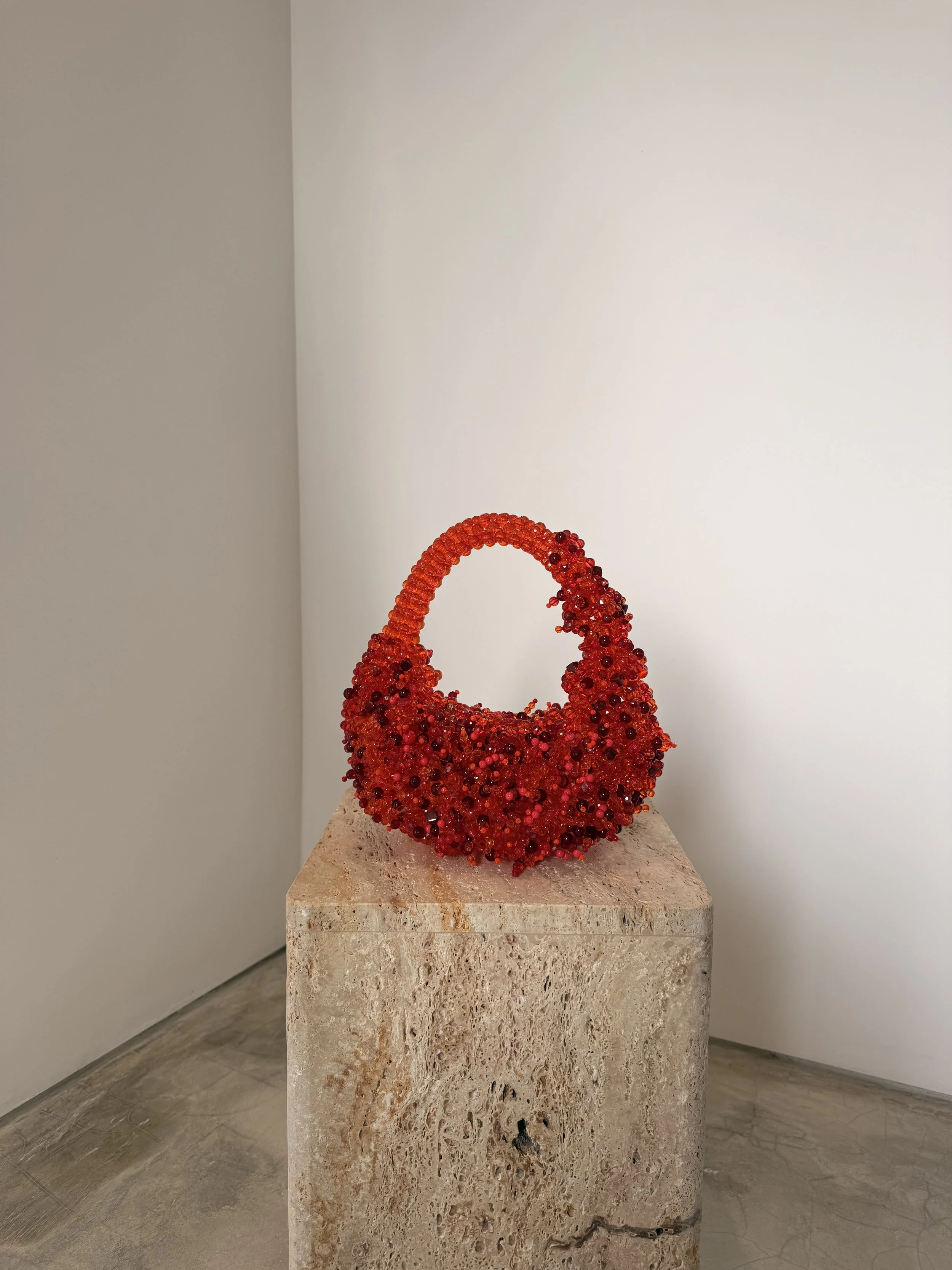 A red handbag sculpture made of small red beads and dark accents, displayed on a beige stone pedestal against a white wall in a gallery setting.
