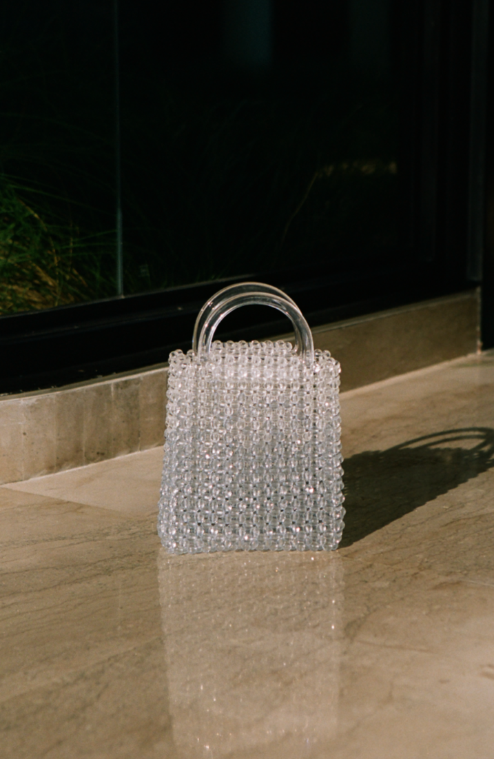 A transparent handbag made of beaded material, resting on a marble floor near a glass window.
