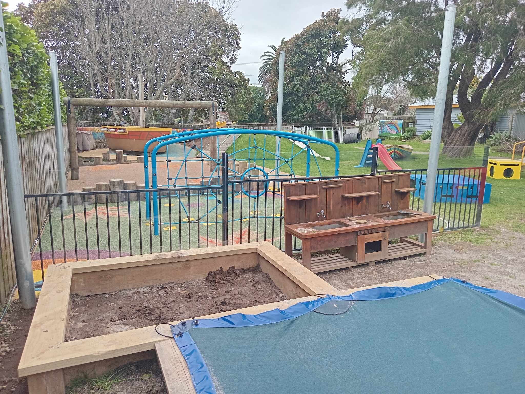 playground with climbing structure, slide, sandbox, and outdoor sinks, surrounded by trees and grass